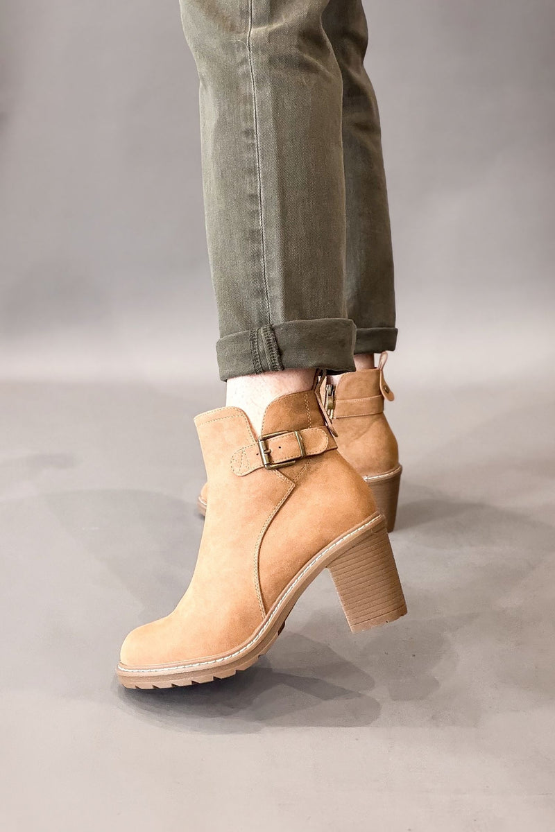 A pair of camel faux suede ankle boots with a block heel and side buckle strap, shown on a model wearing cuffed olive green jeans against a neutral gray studio background.