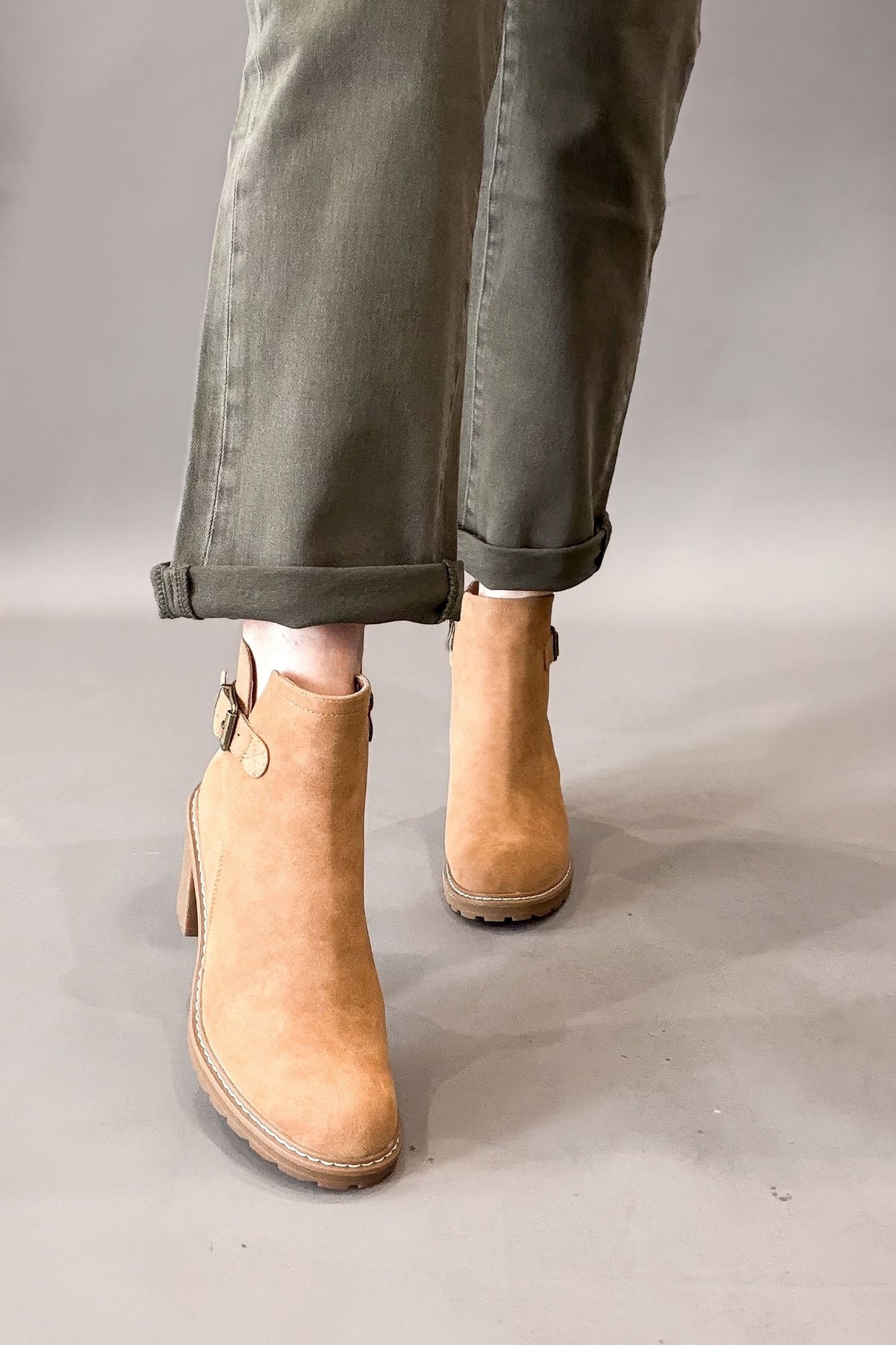 A person wearing camel faux suede ankle boots with side buckle details, paired with cuffed olive green jeans, shown from the knees down against a neutral gray studio background.