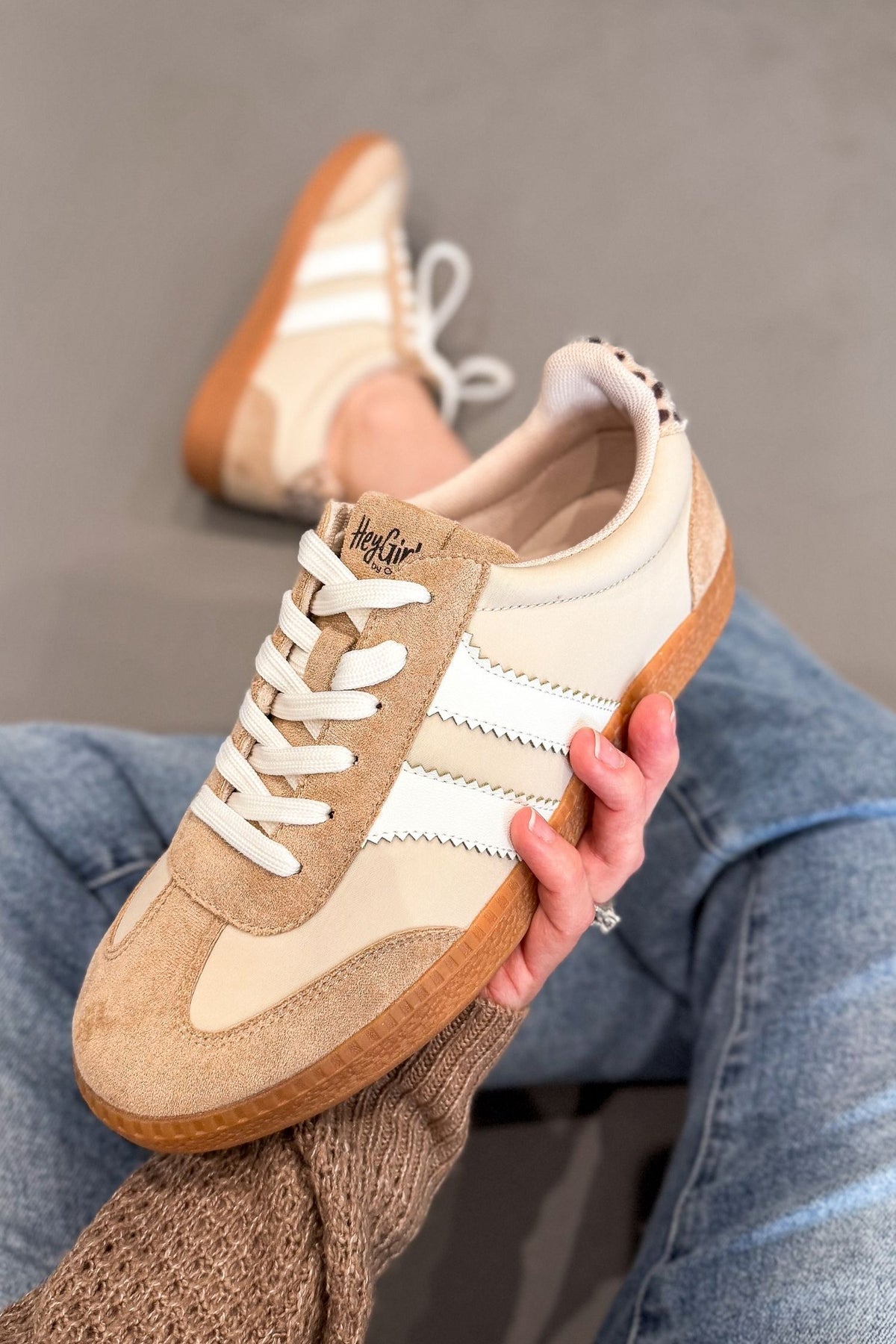A person holds a beige and cream suede sneaker with white laces and white side stripes, while wearing the matching sneaker on their foot. The person is wearing blue jeans and a brown knit sweater, against a gray background.