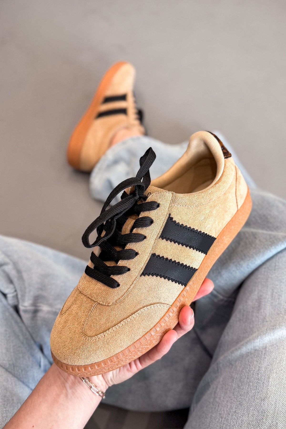 A person holds a beige faux suede sneaker with black stripes and black laces, while wearing the same sneaker with light blue jeans in the background.