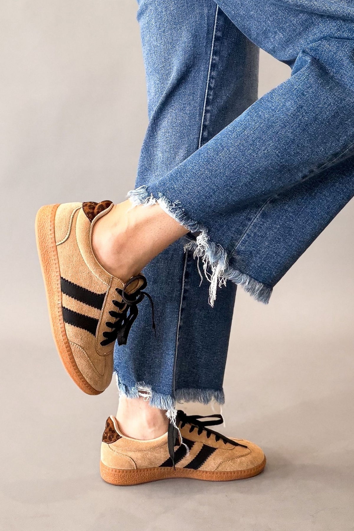 A person wearing blue frayed-hem jeans and tan suede sneakers with black three-stripes, shown from the knees down against a plain light gray studio background.