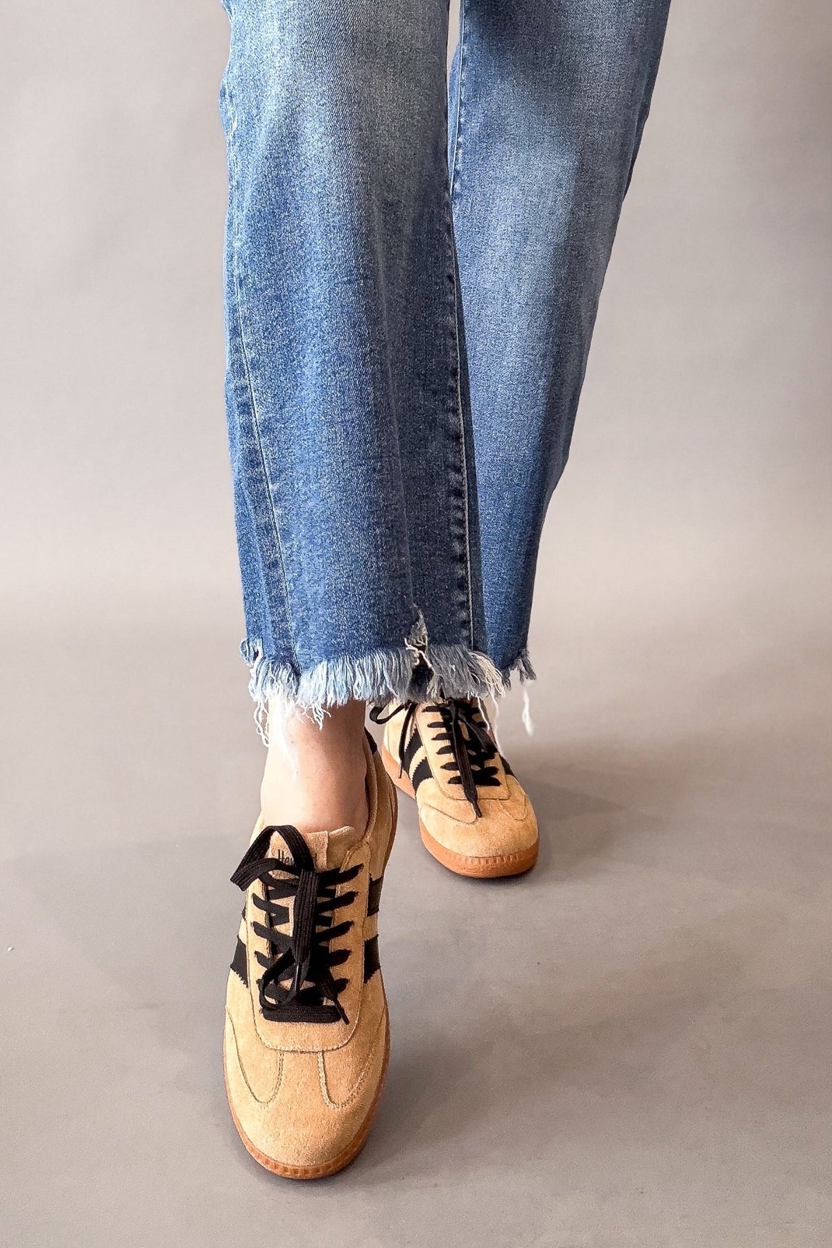 A person wearing light blue frayed-hem jeans and tan suede sneakers with black laces, shown from the knees down against a plain gray studio background.