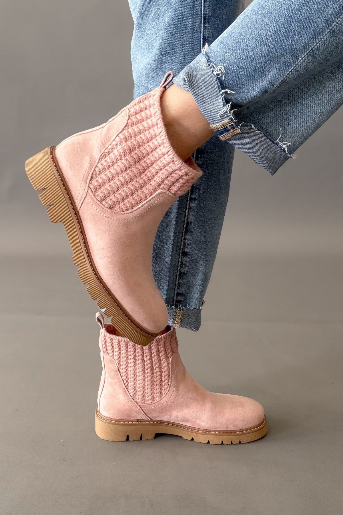 A person wearing light pink ankle boots with knit elastic side panels and tan lug soles, paired with cuffed blue jeans against a neutral gray studio background.