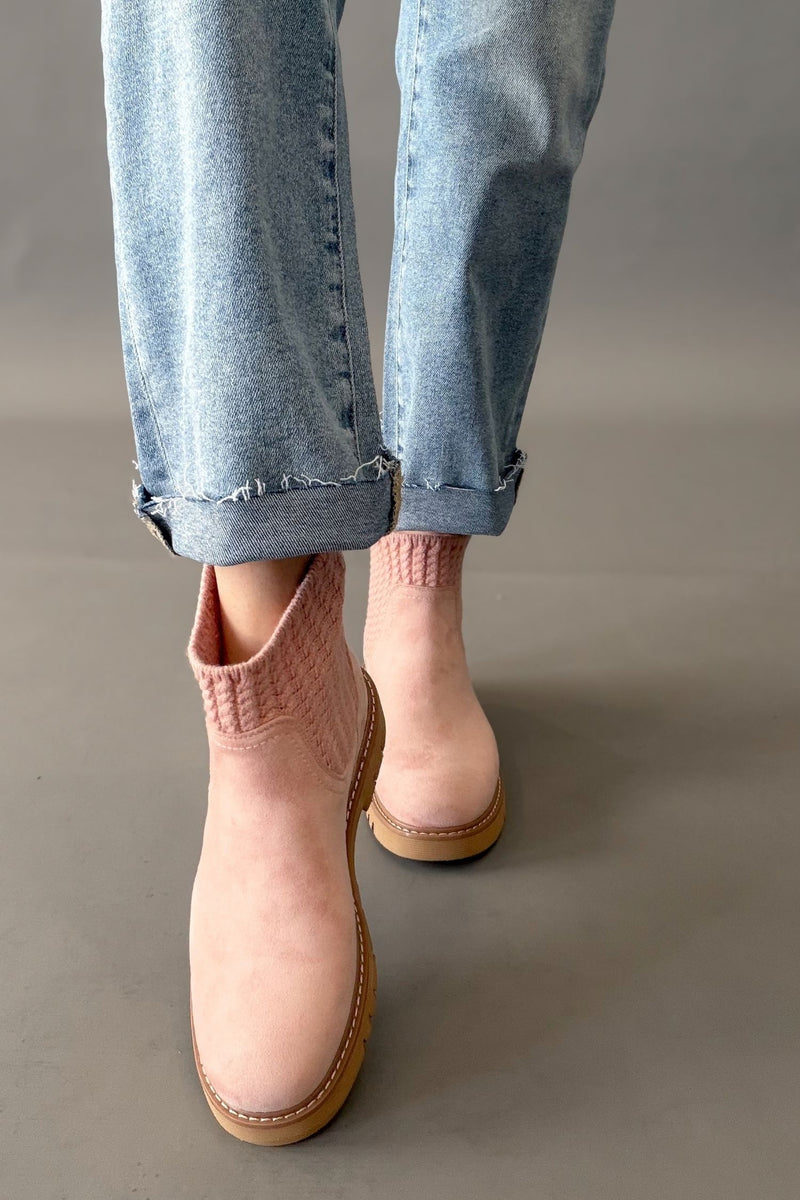A person wearing light blue cuffed jeans and pale pink knit ankle boots with tan soles, standing against a plain gray studio background.