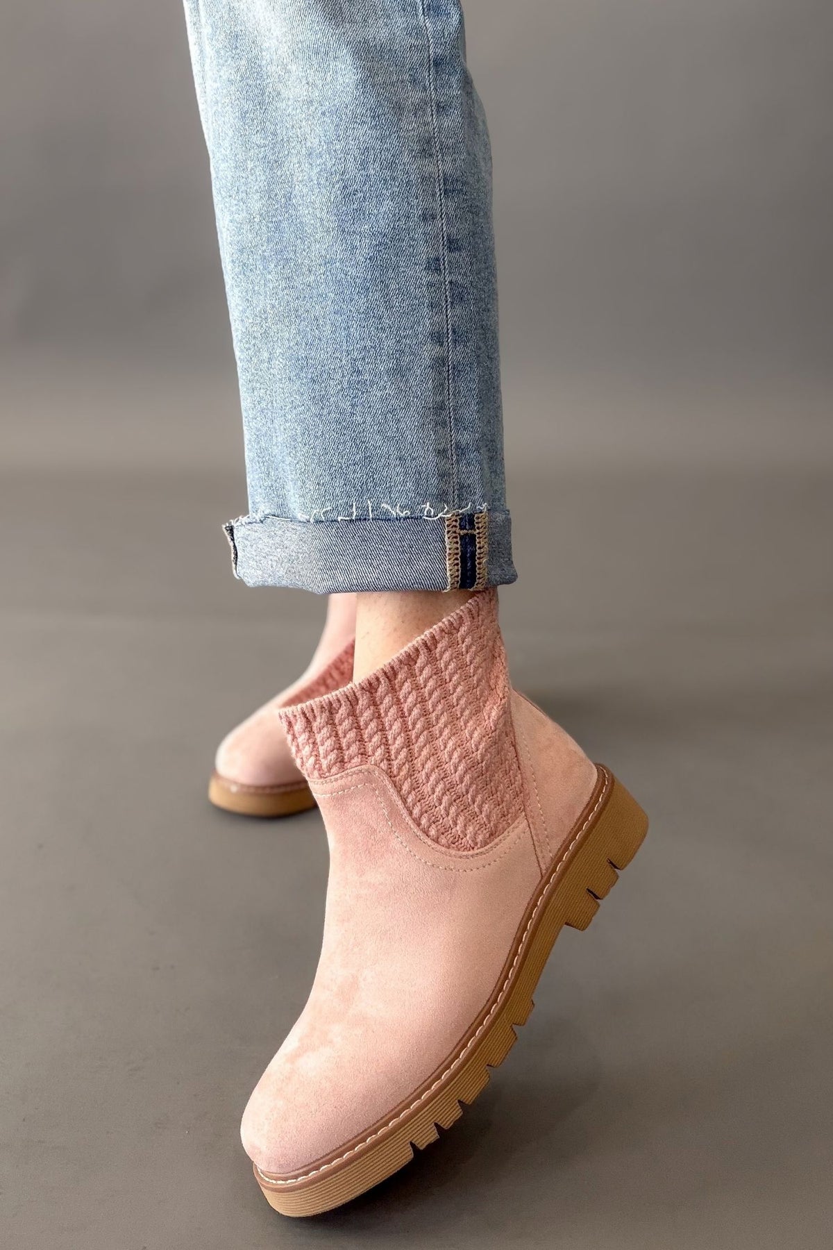 A person wearing light blue jeans and pale pink ankle boots with a knit elastic side panel and tan lug soles, photographed against a smooth gray studio background.