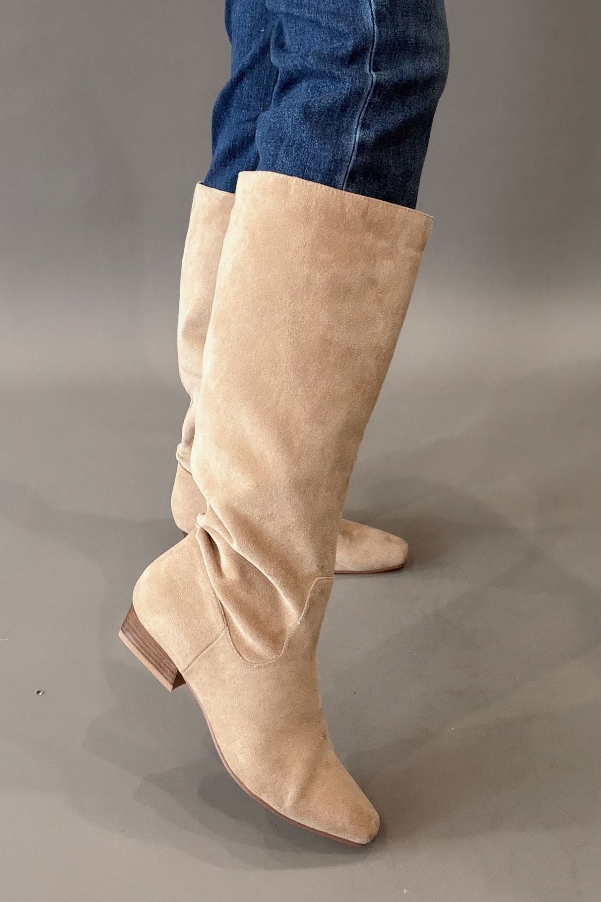 A pair of knee-high camel corkys suede boots worn with blue jeans, shown from a side angle against a neutral gray studio background.