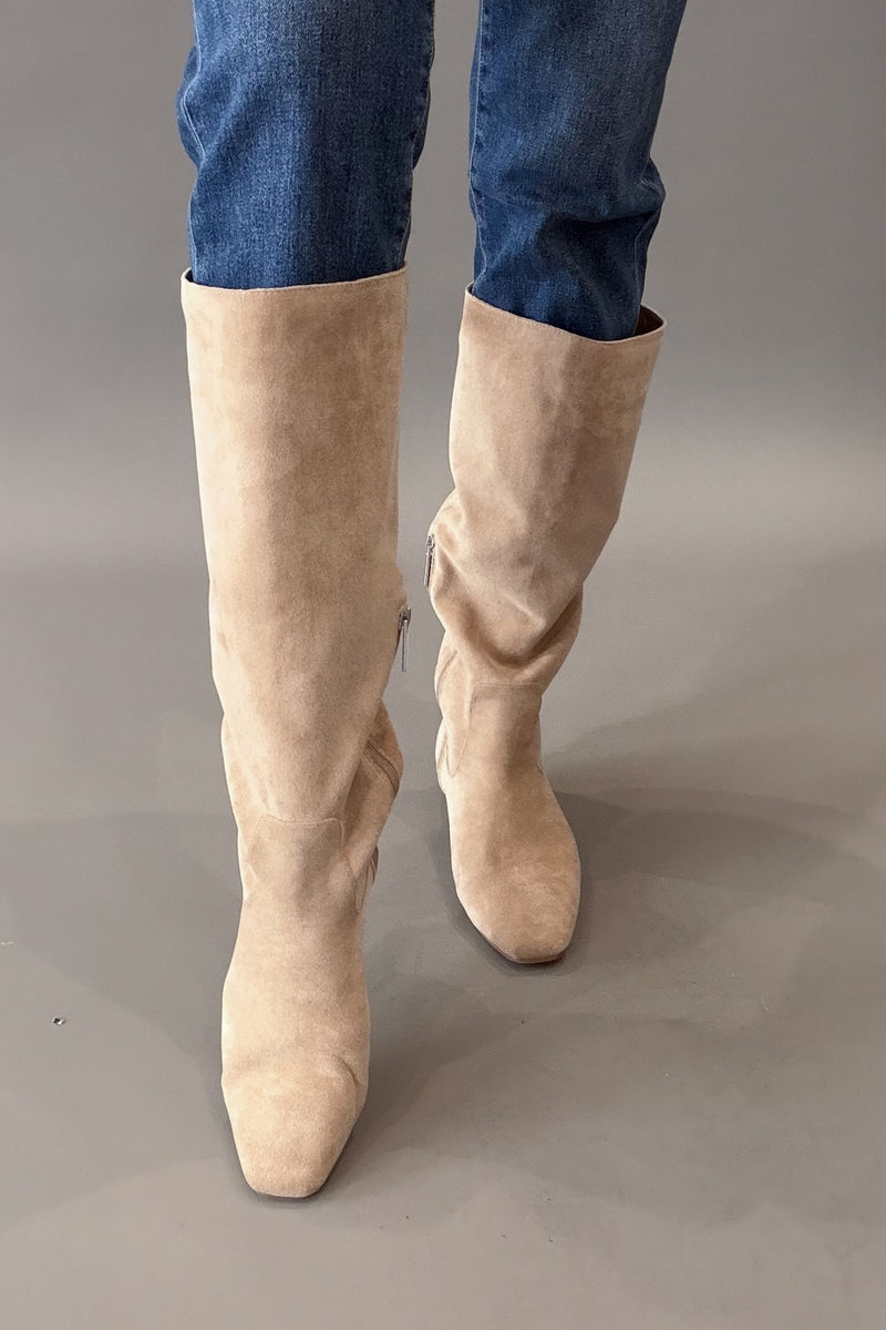 A pair of knee-high camel suede boots worn with blue jeans against a neutral gray studio background.