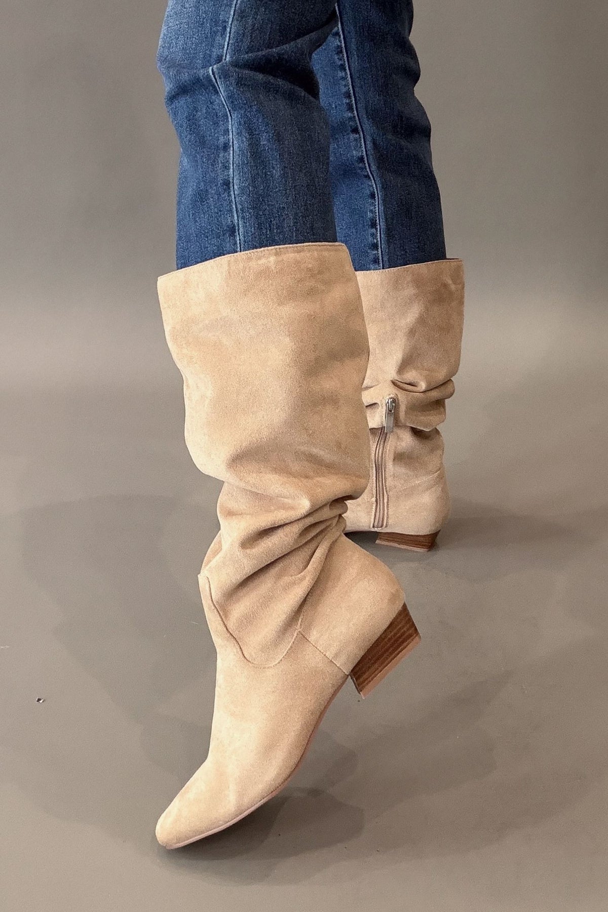 A pair of camel suede slouchy knee-high boots with pointed toes and stacked block heels, shown on a model wearing blue jeans against a neutral gray studio background.