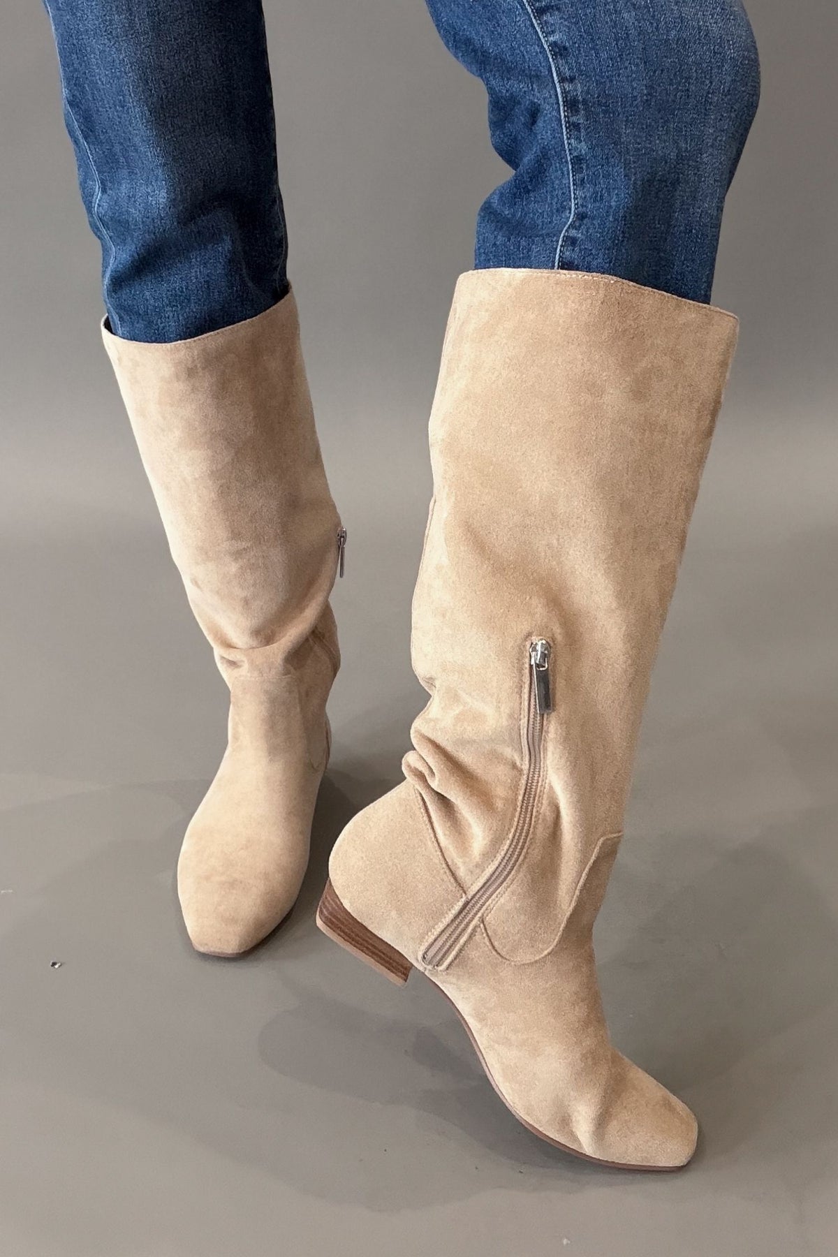 A pair of knee-high camel suede boots with a side zipper and low block heel, shown on a model wearing blue jeans against a gray studio background.