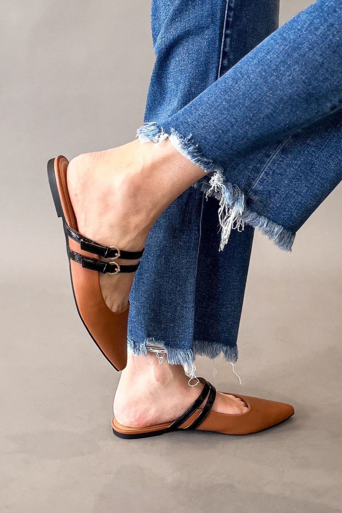 A person wearing congnac pointed-toe flat mules with black double straps and gold buckles, paired with frayed blue jeans, against a plain gray studio background.