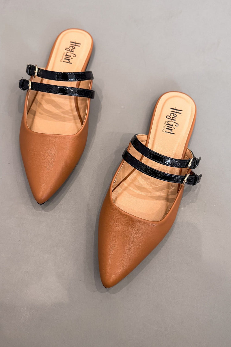 A pair of pointed-toe flat mule mules in cognac with black double straps and gold-tone buckle details, displayed on a neutral gray background.