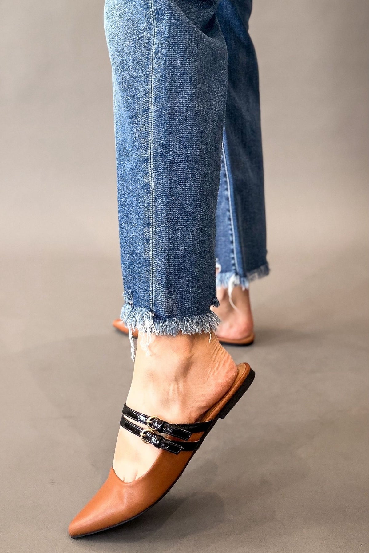 A person wearing blue frayed-hem jeans and cognac pointed-toe mule flats with black double straps, shown against a plain gray studio background.