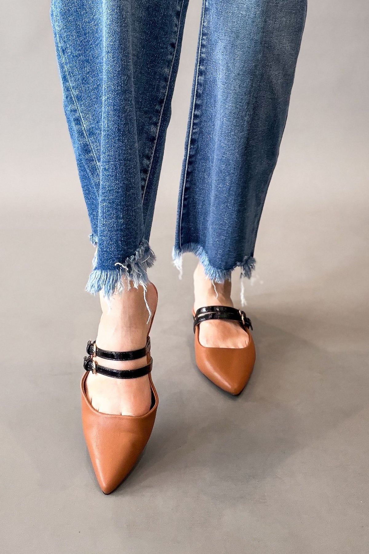A person wearing blue frayed-hem jeans and cognac pointed-toe mule heels with black double straps, standing against a plain gray studio background.