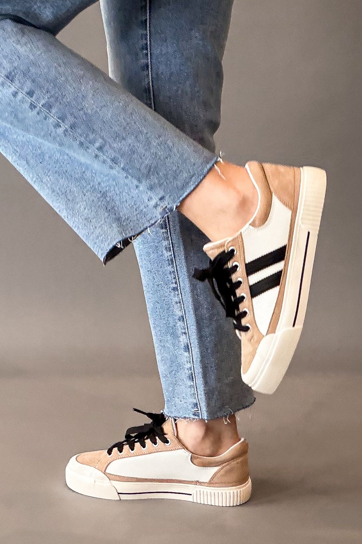 A person wearing light blue jeans and beige and white platform sneakers with black stripes and black laces, shown from the knees down against a plain gray studio background.