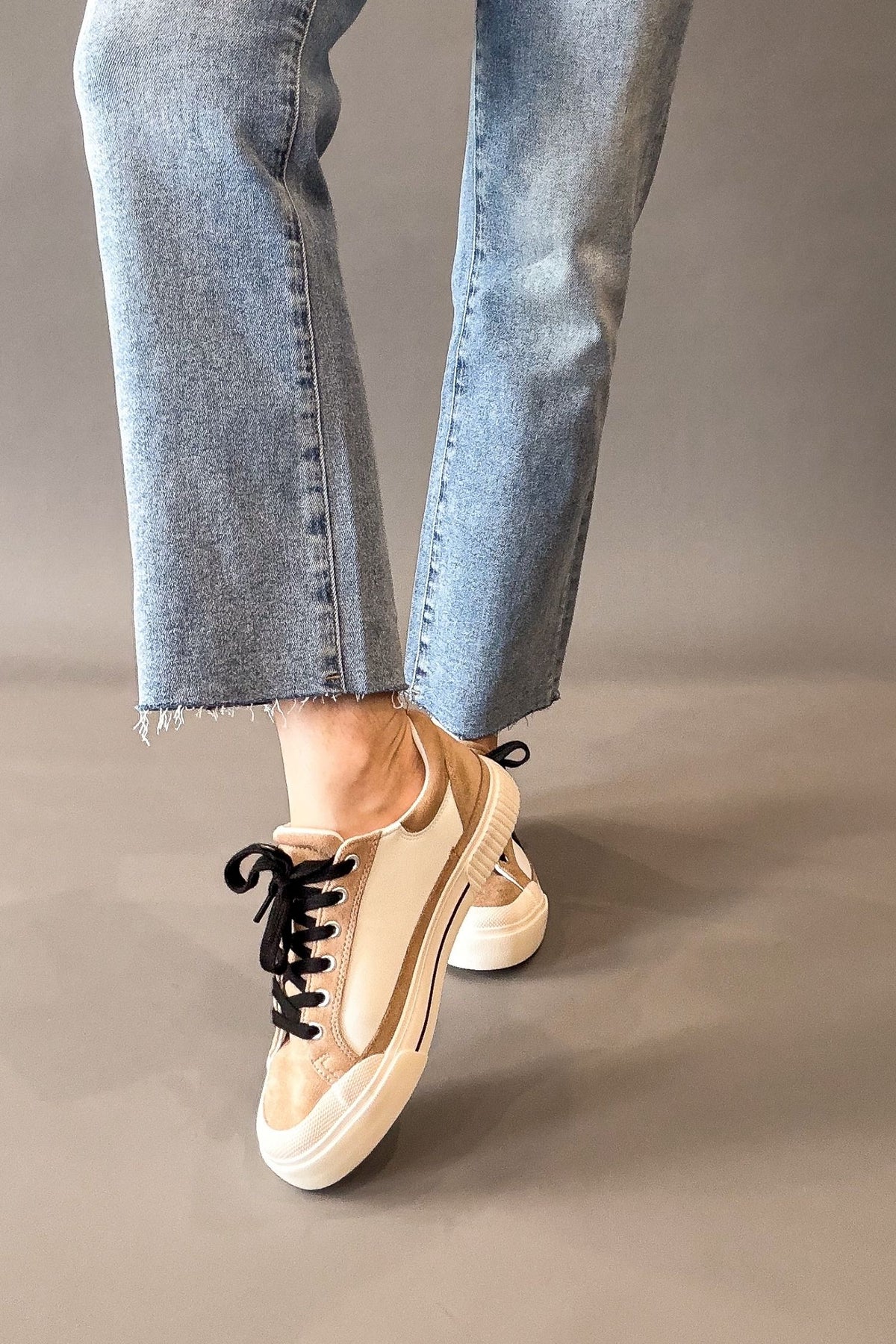 A person wearing light blue cropped jeans and beige low-top platform sneakers with black laces, shown from the calves down against a plain taupe studio background.