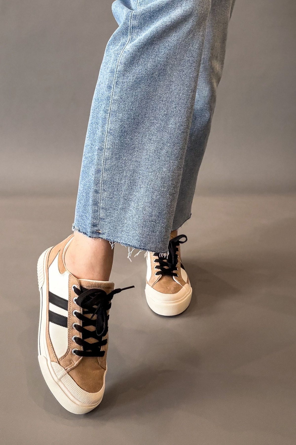 A person wearing light blue frayed-hem jeans and tan low-top sneakers with black stripes, black laces, and off-white soles, standing against a plain gray studio background.