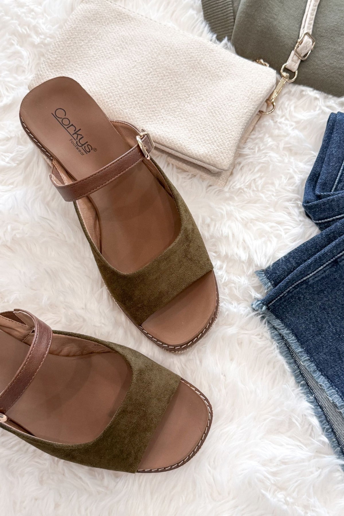 A pair of women's wedge sandals in olive moss faux suede with brown straps, styled in a flat lay on a white faux fur rug alongside a cream clutch, blue jeans, and a ivory bag.