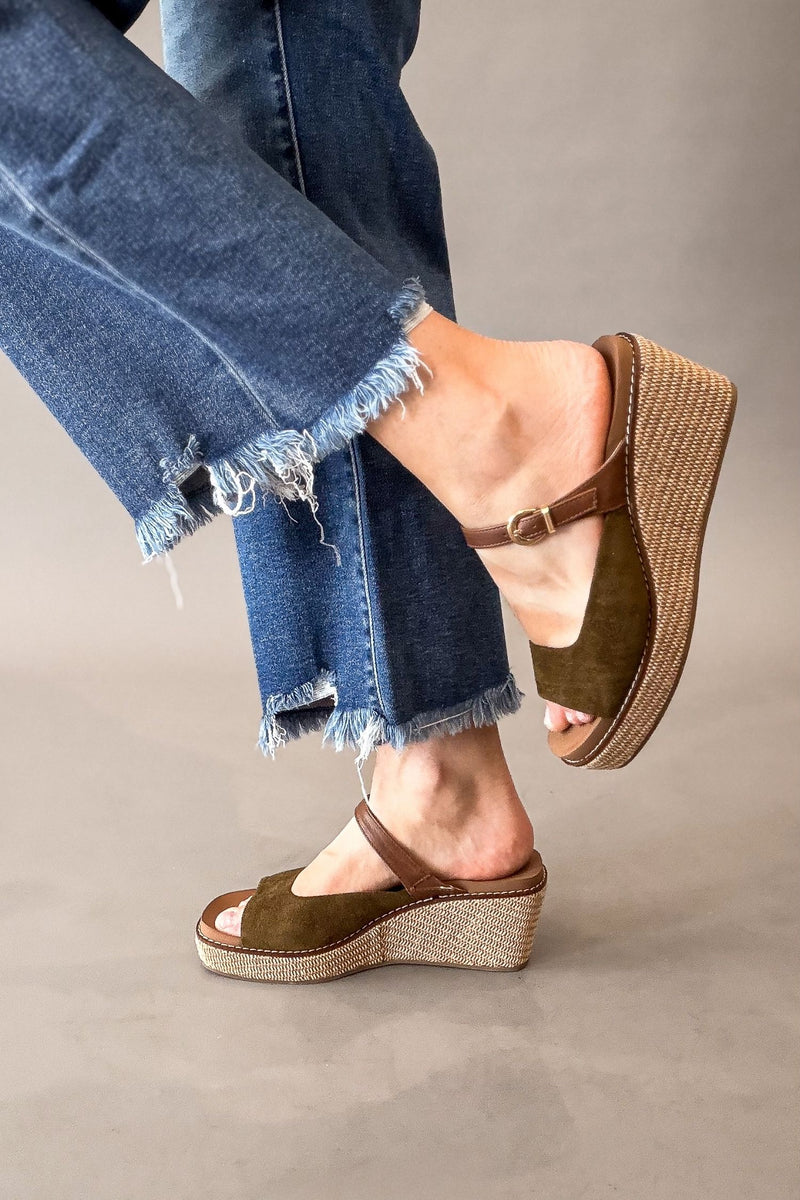 A person wearing olive green wedge sandals with braided jute soles and blue frayed-hem jeans, shown from the calves down against a plain gray studio background.