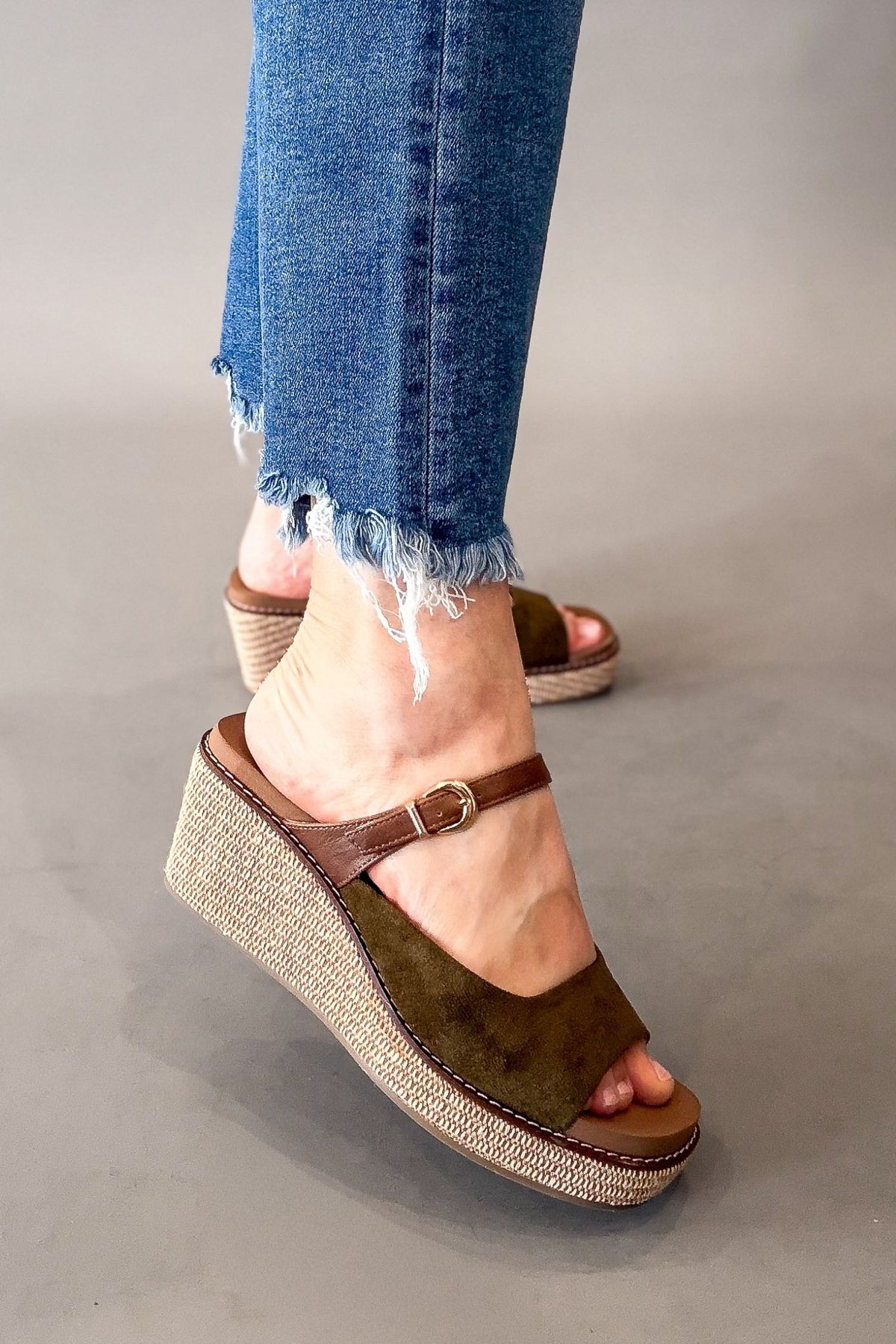 A person wearing moss green wedge sandals with brown straps and frayed blue jeans, shown from the calves down on a plain gray studio background.