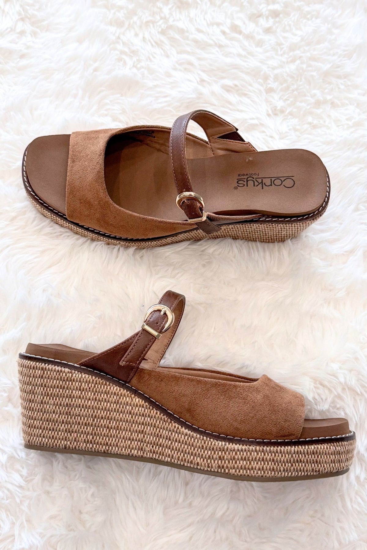 A pair of women's wedge sandals in brown faux suede and leather with woven jute-wrapped wedge heels, displayed on a white faux fur background. One sandal is shown from a top-down view and the other from a side profile view, both featuring a small gold buckle on the ankle strap.