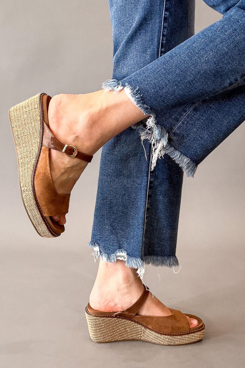 A person wearing chestnut faux suede wedge sandals with braided jute-wrapped platform soles, paired with frayed blue jeans, shown against a neutral gray studio background.