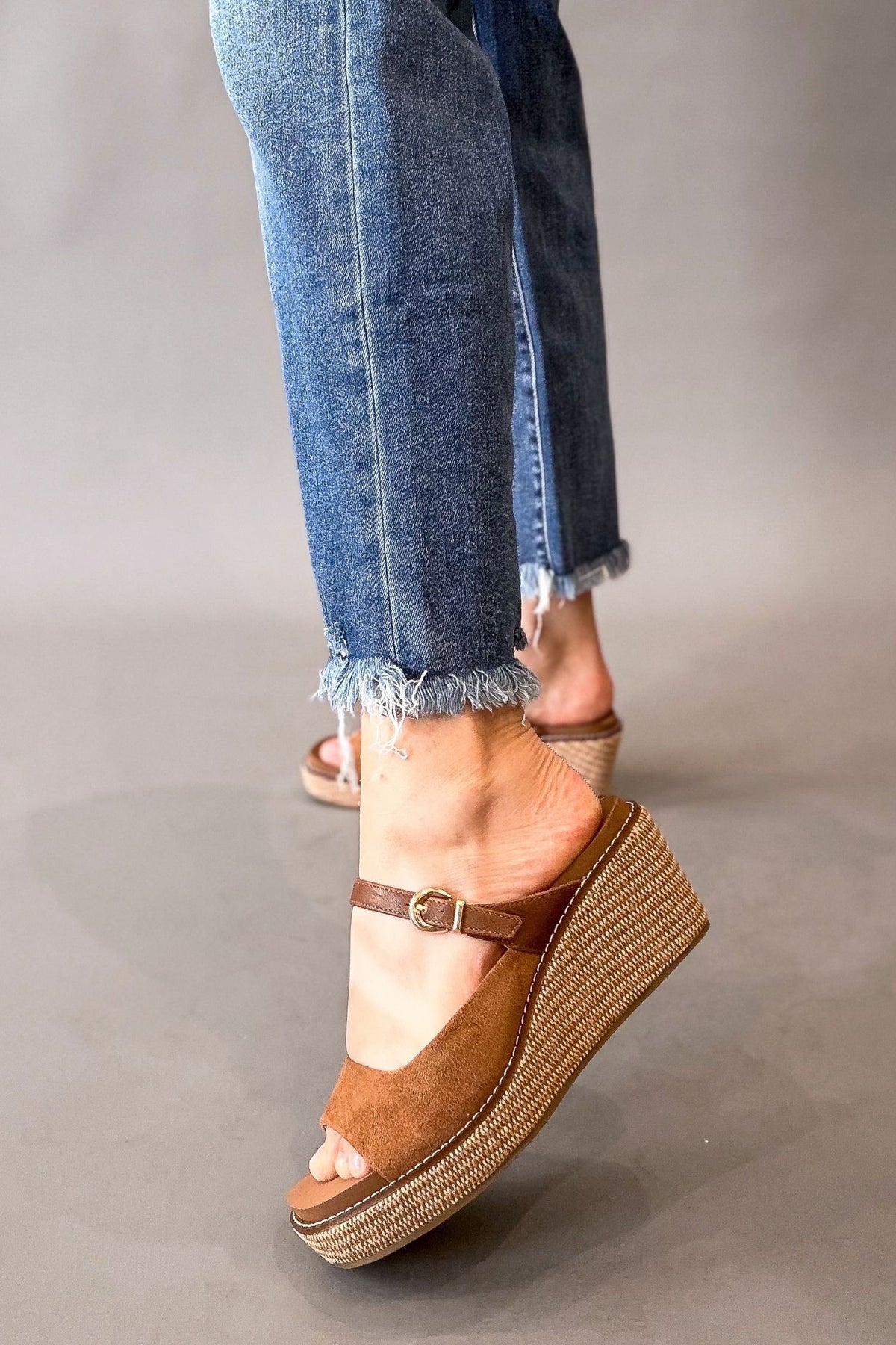 A person wearing chestnut suede wedge sandals with jute-style woven platforms and blue frayed-hem jeans, shown from the knees down against a plain gray studio background.