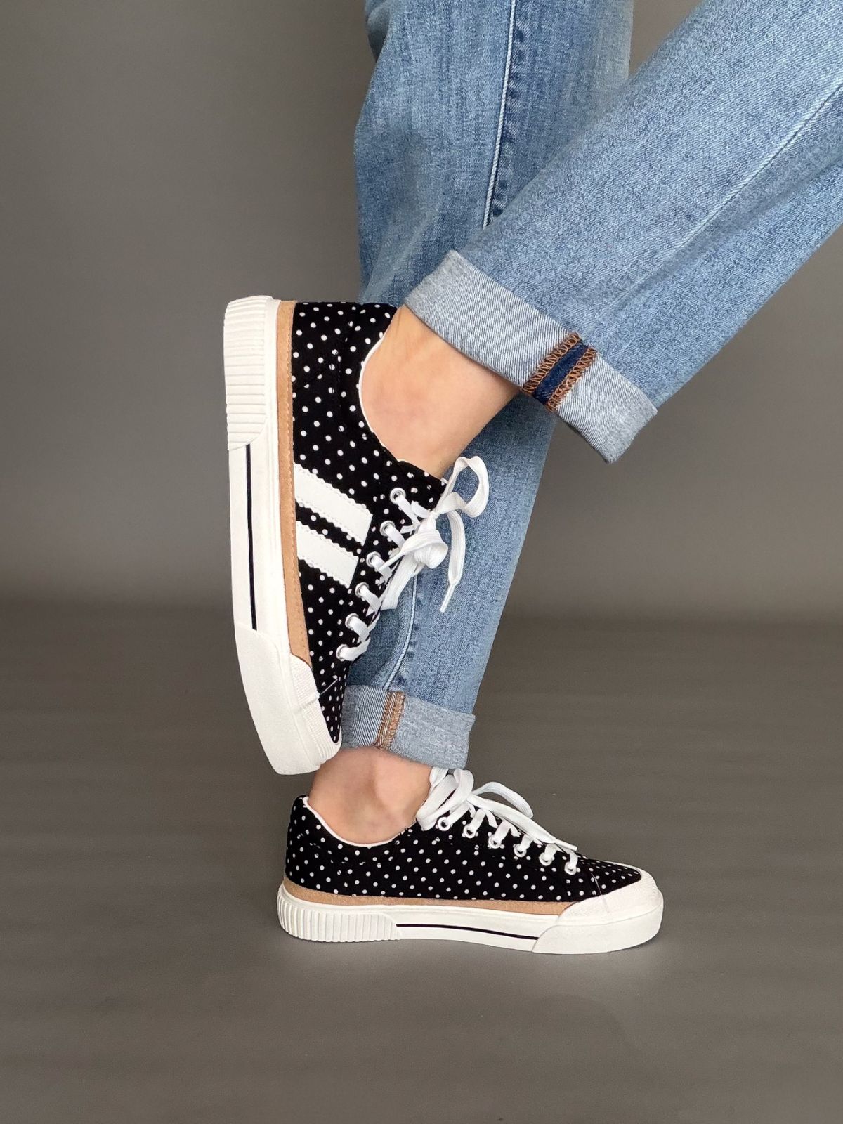 woman wearing corkys Black polka dot sneakers with white laces worn with blue jeans on a gray background