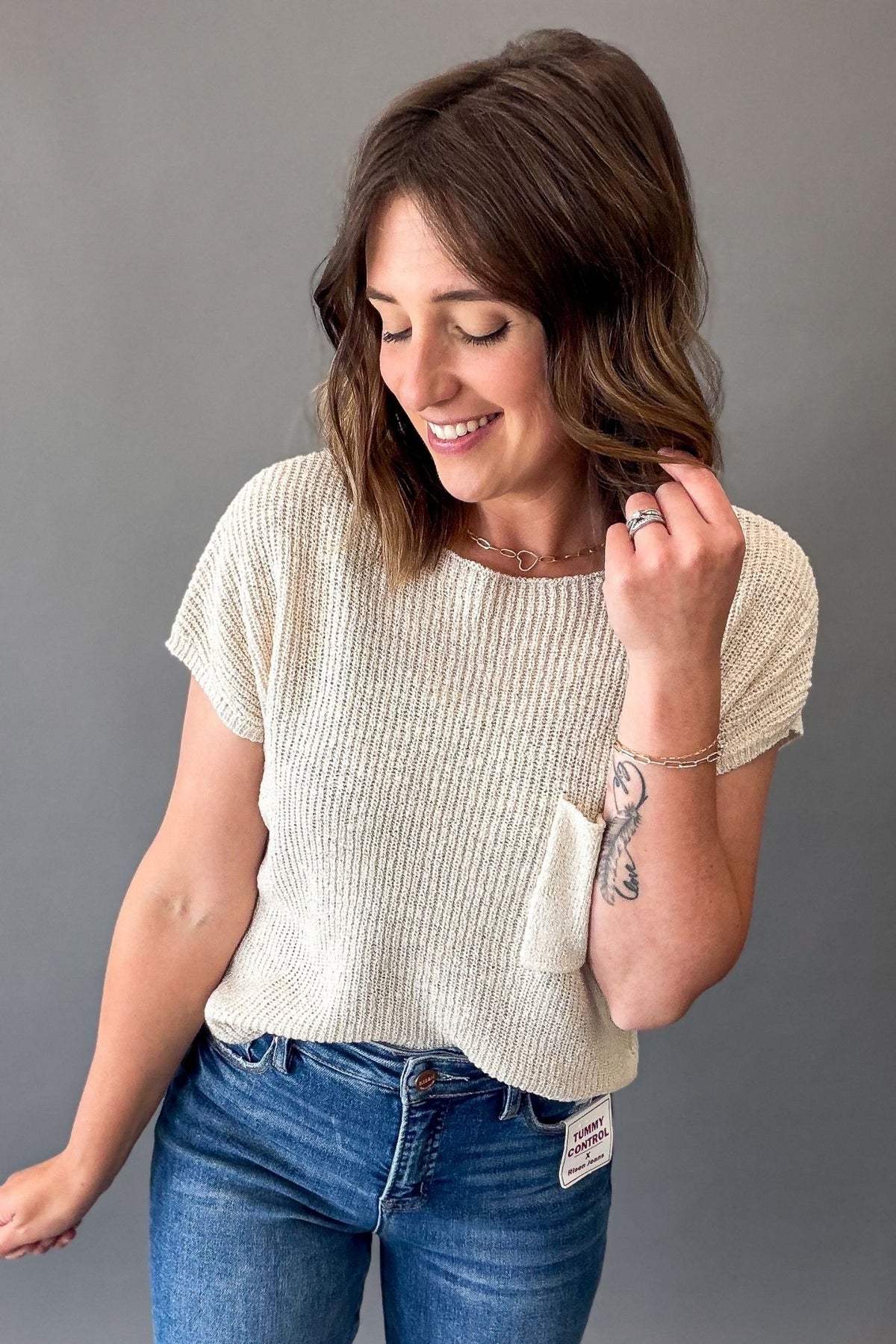 A woman wearing a cream-colored short-sleeve ribbed knit top and blue jeans, standing against a solid gray background.