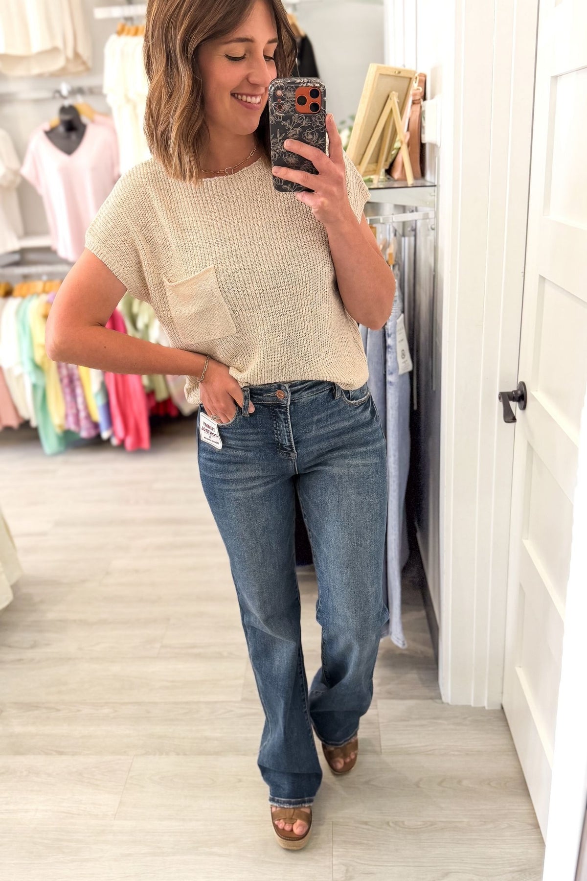A woman takes a mirror selfie in a clothing boutique, wearing a cream short-sleeve knit top and blue risen jeans.