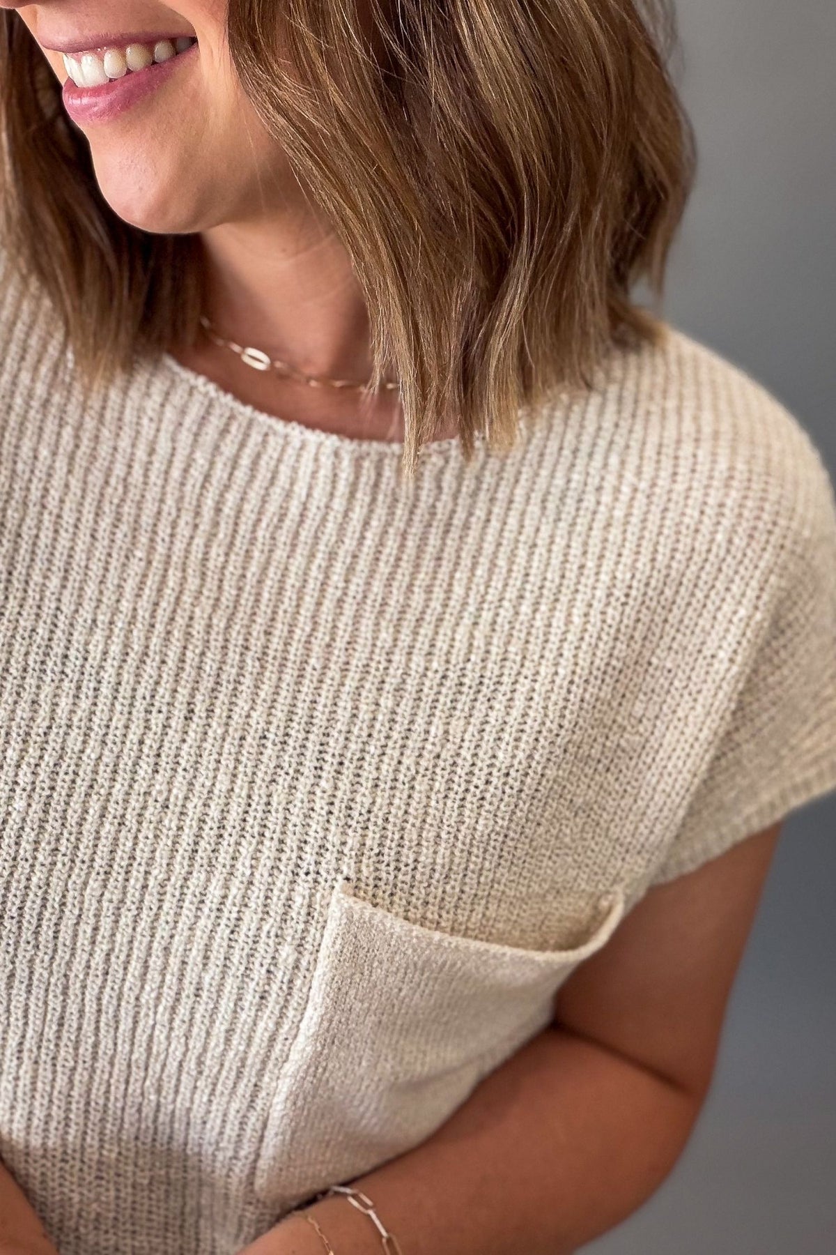 Close-up of a woman wearing a short-sleeve ribbed knit top in a light beige or cream color, featuring a chest pocket and delicate gold jewelry.