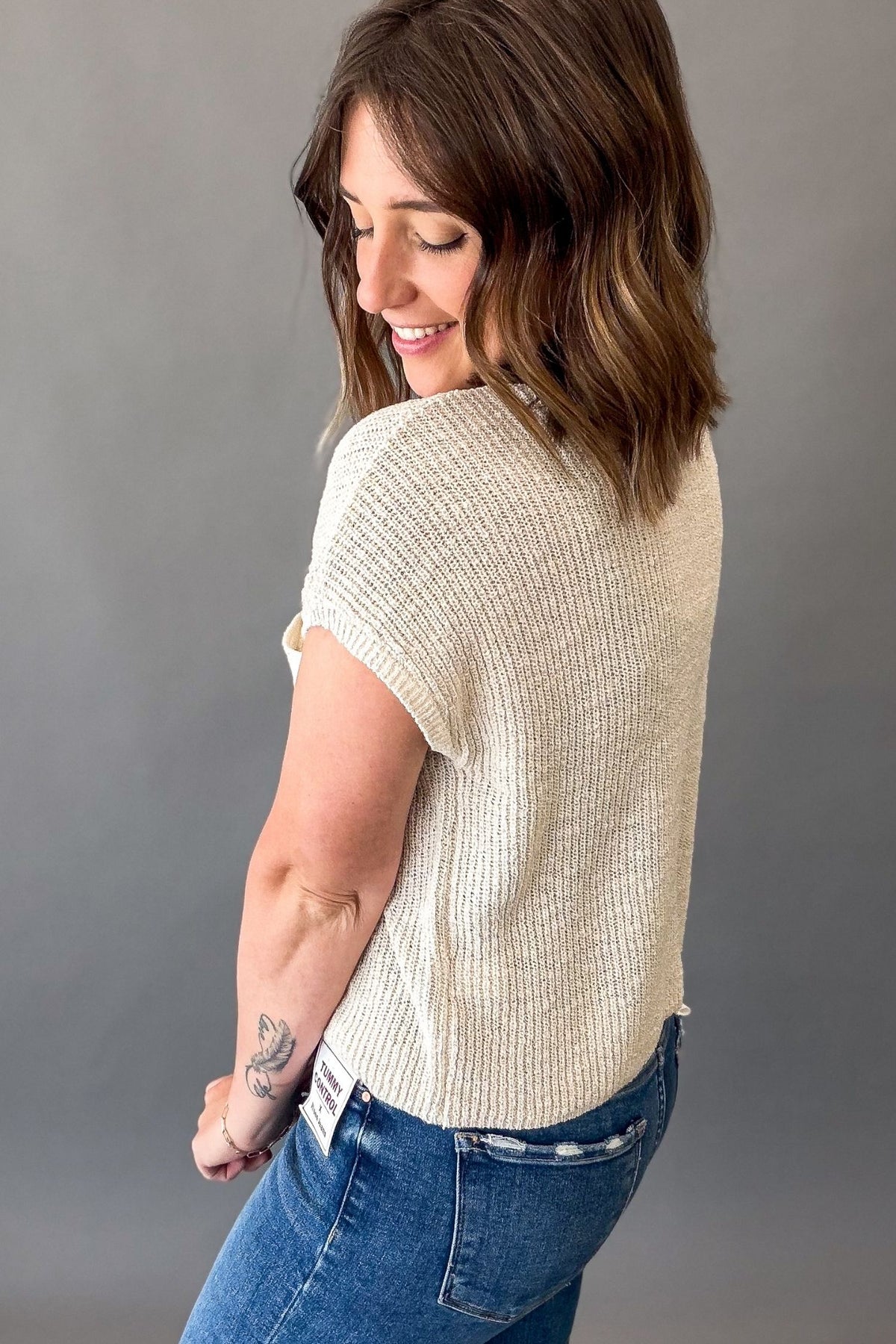 A woman wearing a short-sleeve cream-colored textured knit top and blue jeans, photographed from the back against a solid gray studio background.
