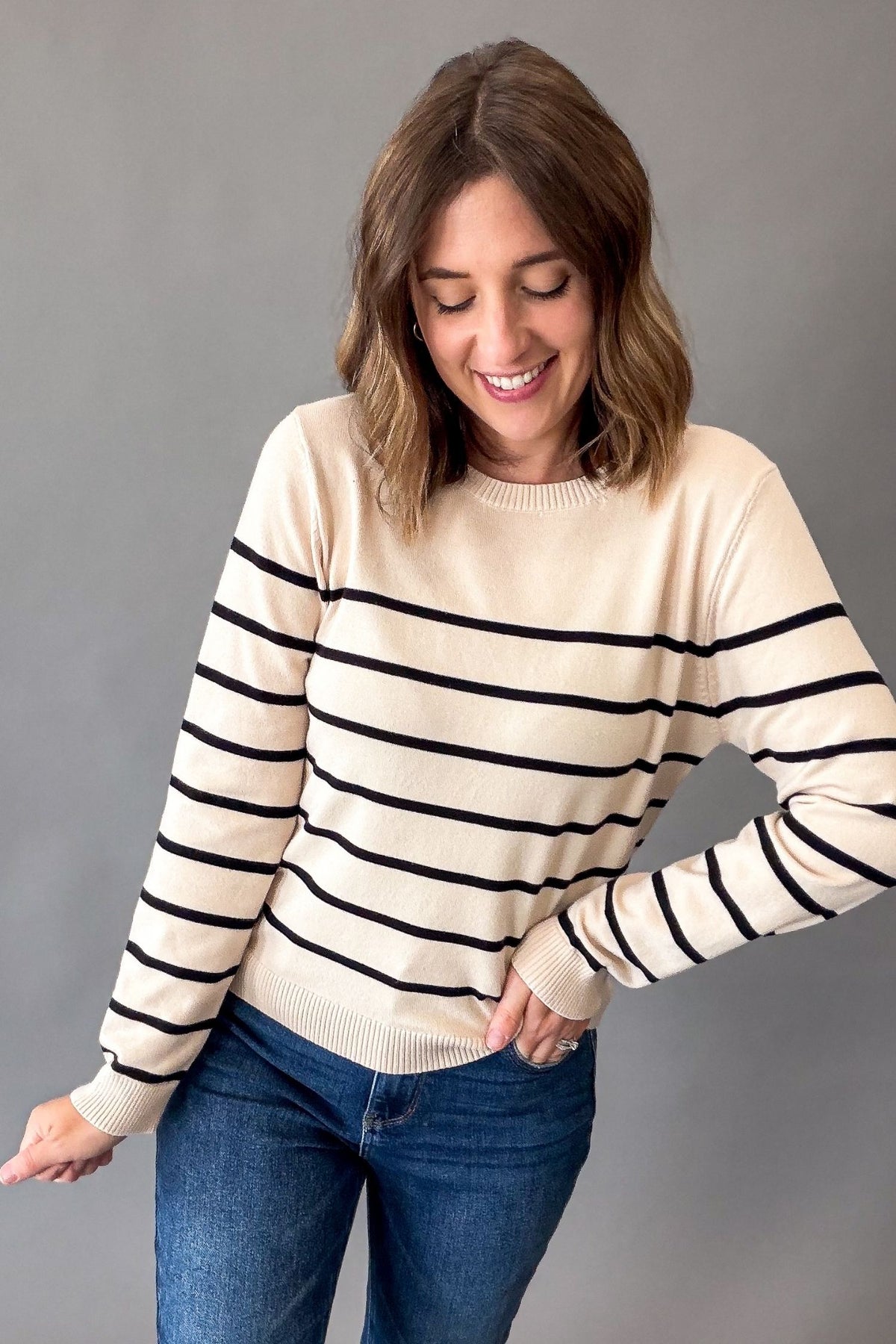 A woman wearing a cream-colored knit sweater with black horizontal stripes and blue jeans, standing against a solid gray studio background.