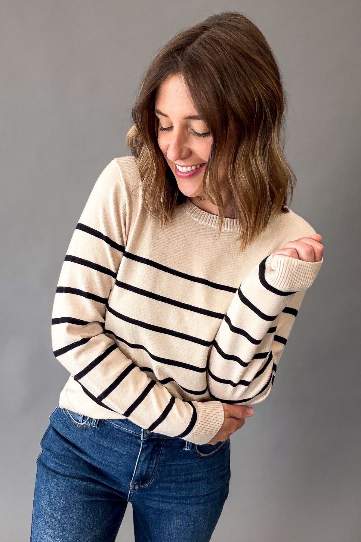 A woman wearing a cream-colored knit sweater with black horizontal stripes and blue jeans, posing against a solid gray studio background.