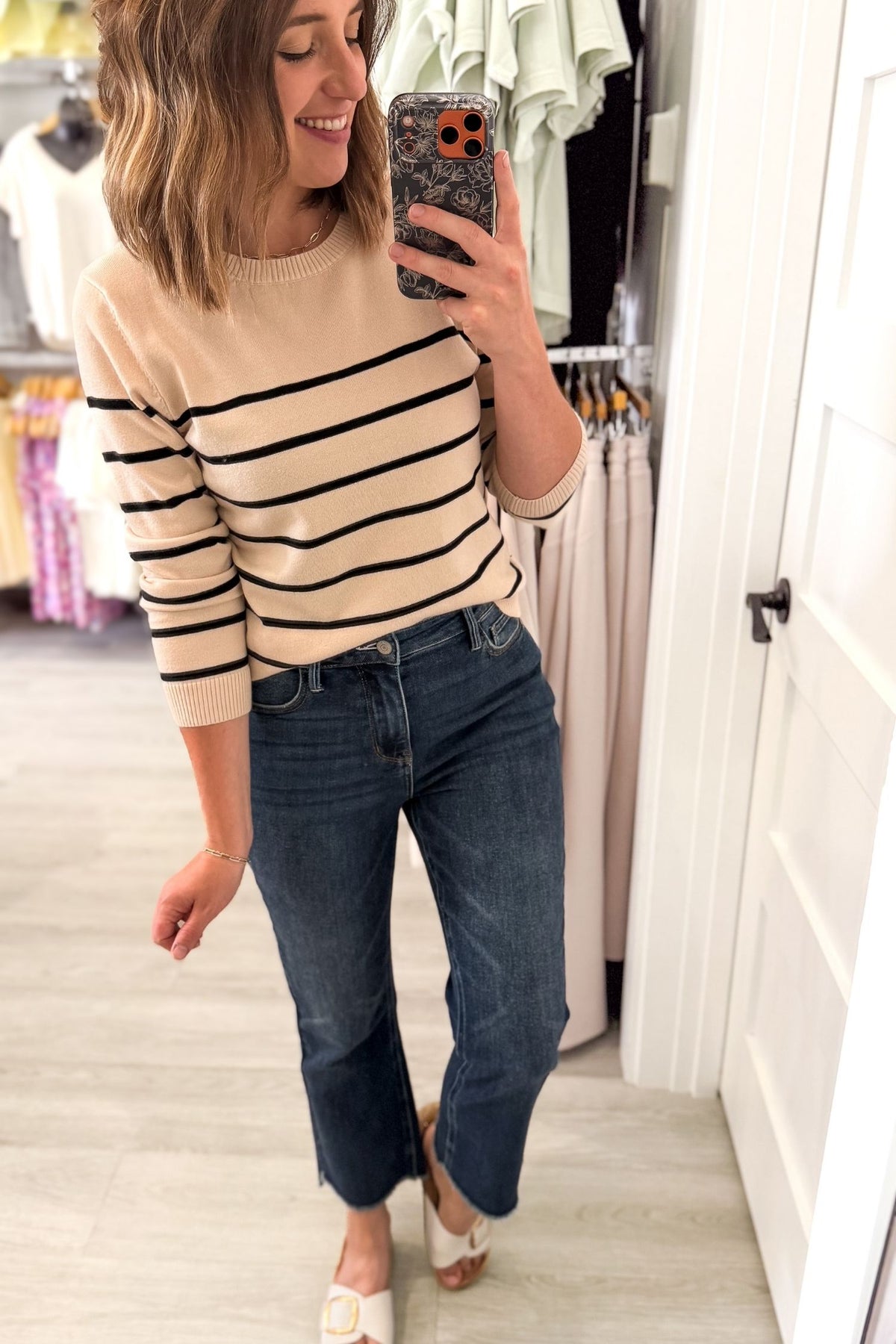 A woman takes a mirror selfie in a clothing store, wearing a beige crewneck sweater with black horizontal stripes, dark blue cropped jeans, and white slide sandals.