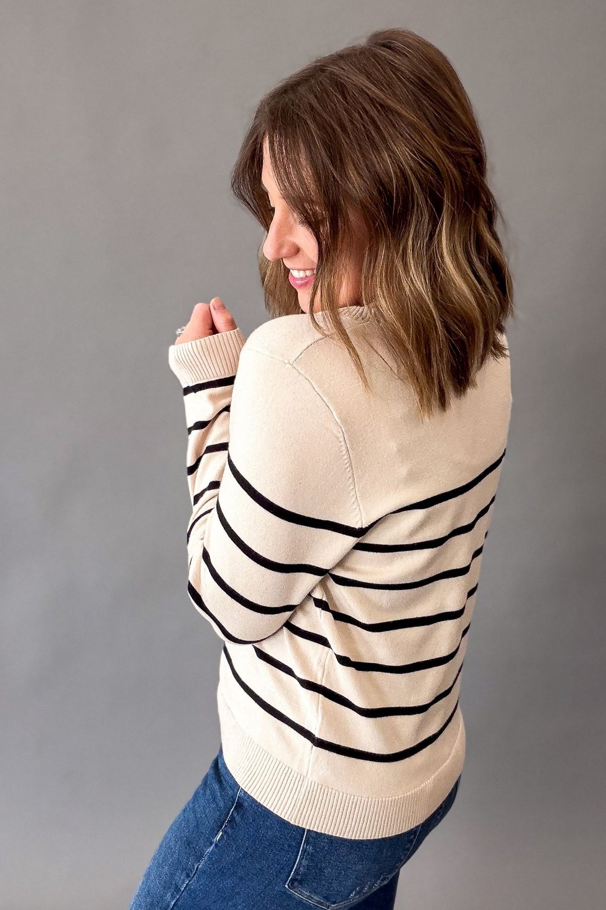 A woman with shoulder-length brown hair is shown from the back wearing a cream knit sweater with horizontal black stripes and blue jeans, posing against a solid gray studio background.