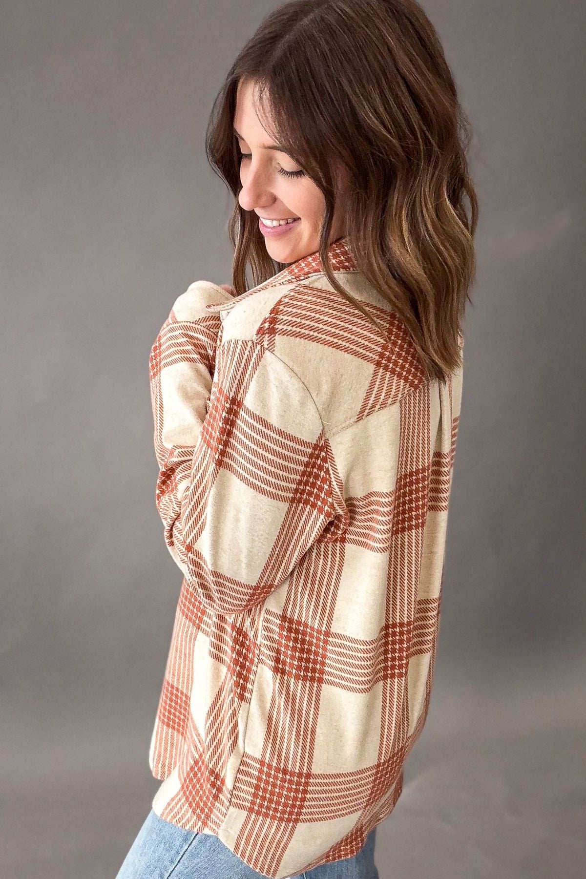 A woman wearing a cream-colored long-sleeve plaid flannel shirt with rust-orange and brown stripes, shown from a side-back angle against a neutral gray studio background.