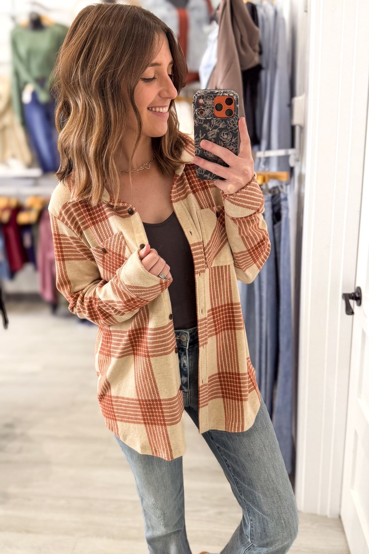 A woman takes a mirror selfie in a clothing store while wearing a beige and rust-orange plaid button-up shirt over a black top and blue jeans.