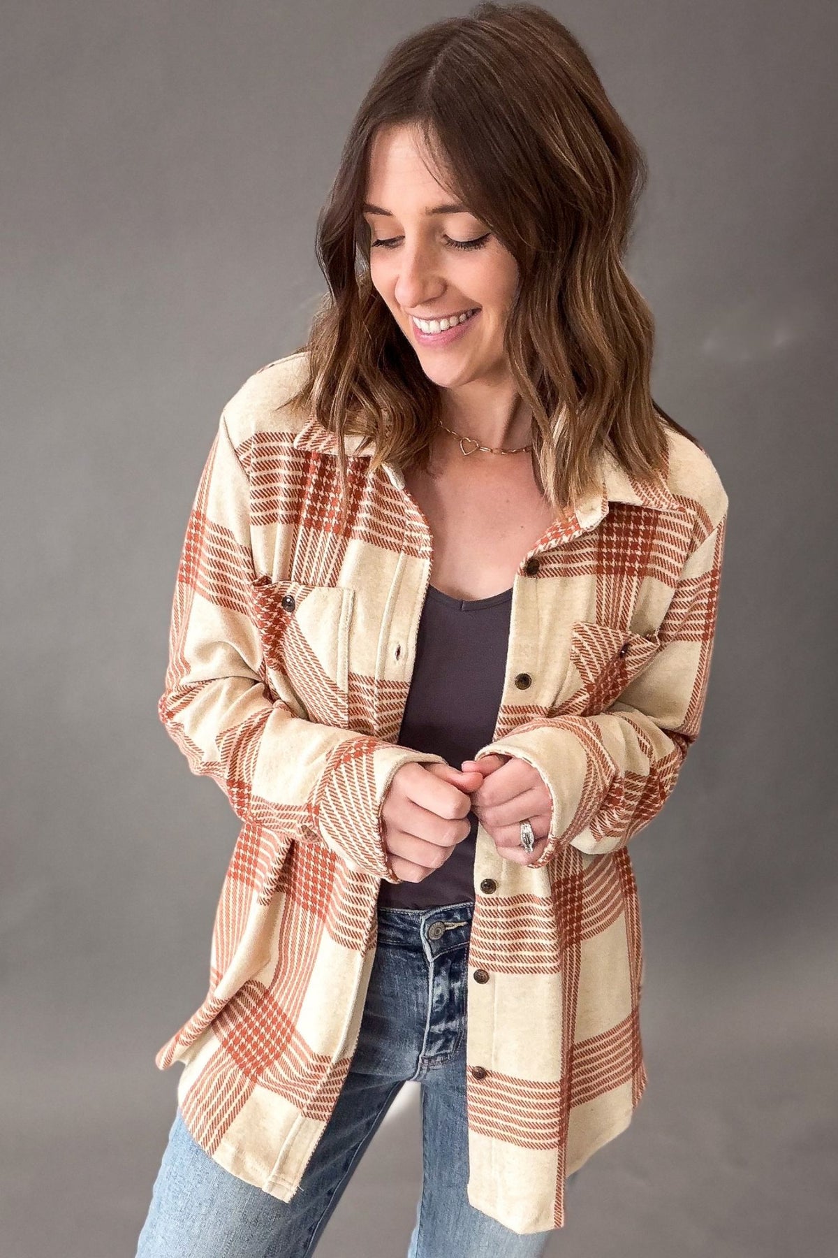 A woman wearing a cream and rust-orange plaid button-up shirt over a dark top and blue jeans, standing against a neutral gray studio background.
