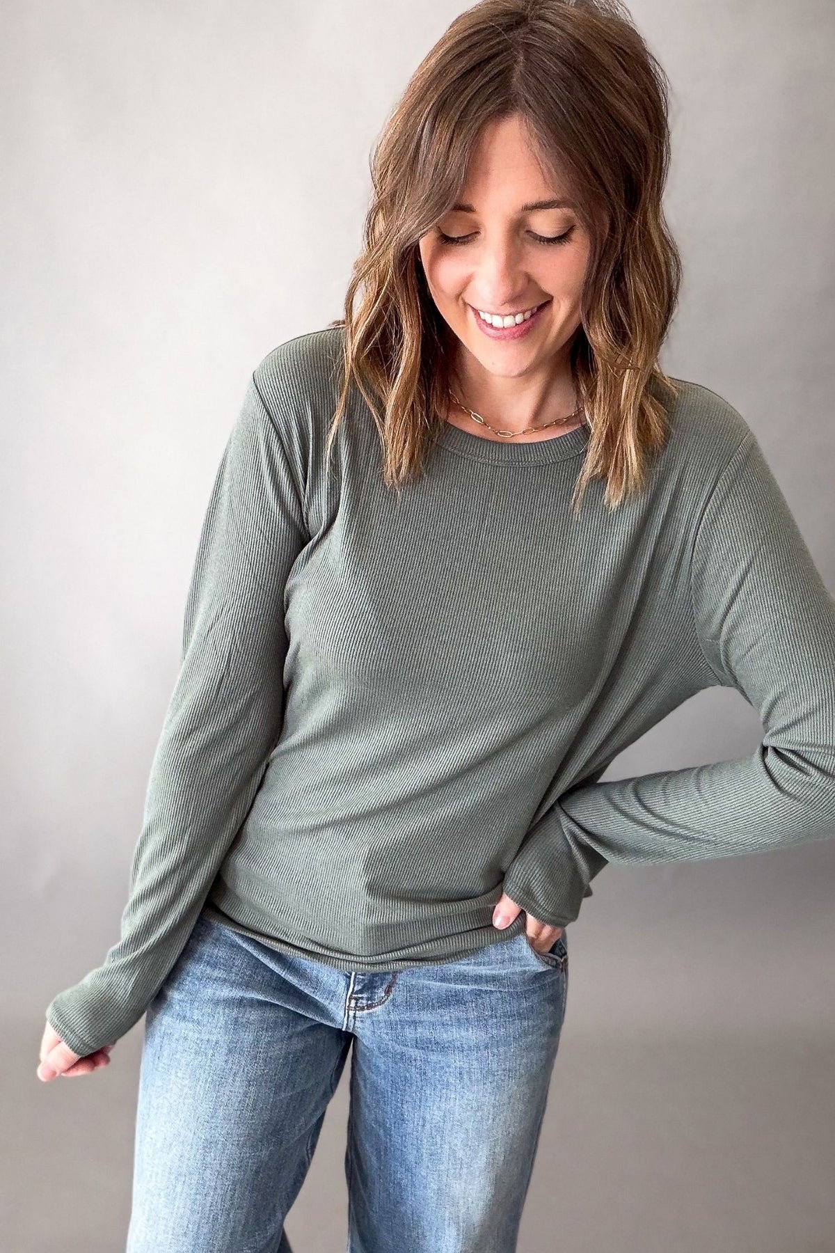 A woman wearing a muted sage green long-sleeve crewneck top and blue jeans, standing against a neutral gray studio background.