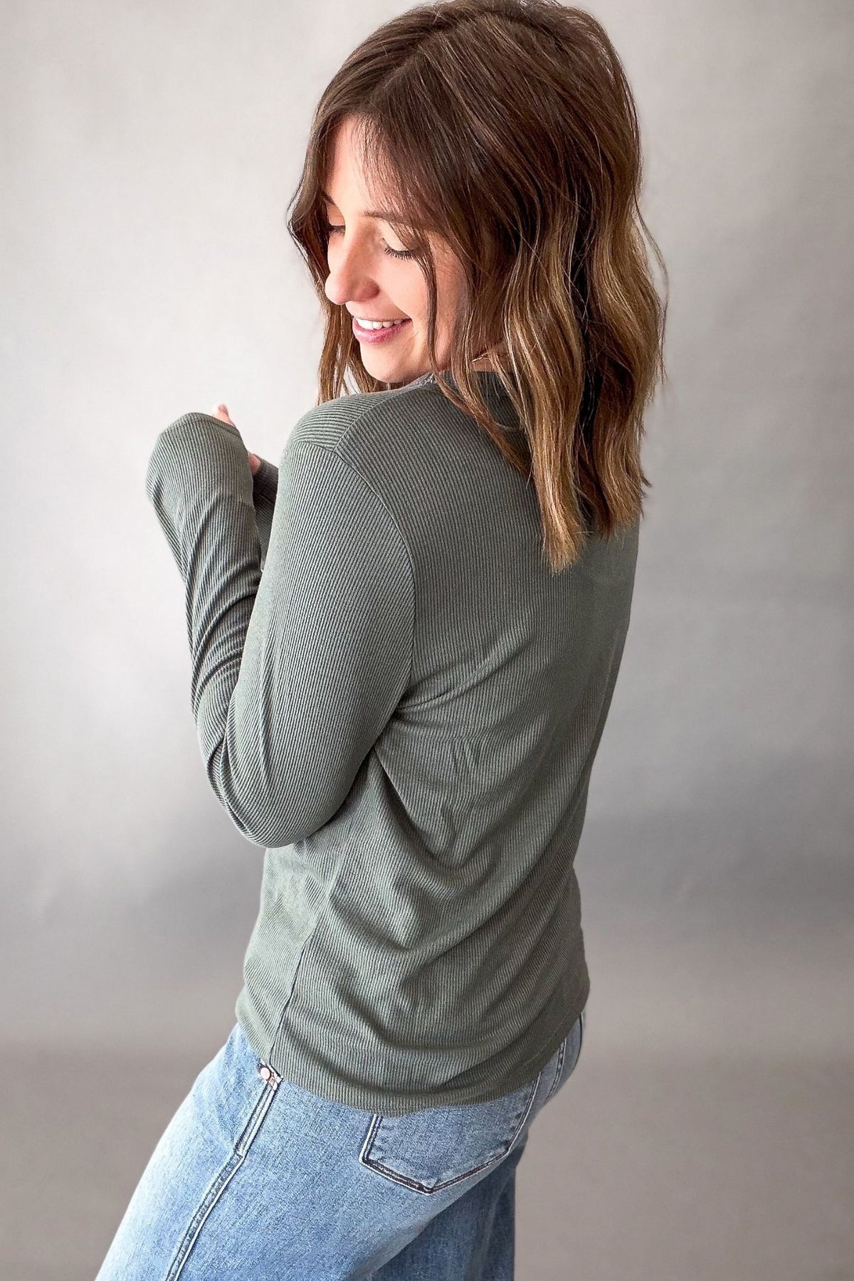 A woman wearing a ribbed long-sleeve turtleneck top in sage green, paired with light blue jeans, photographed against a neutral gray studio background.