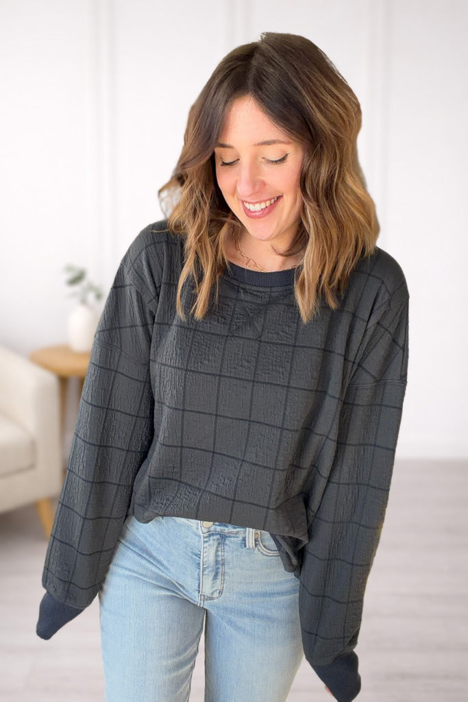 Woman wearing a dark gray checkered sweater and light blue jeans indoors.