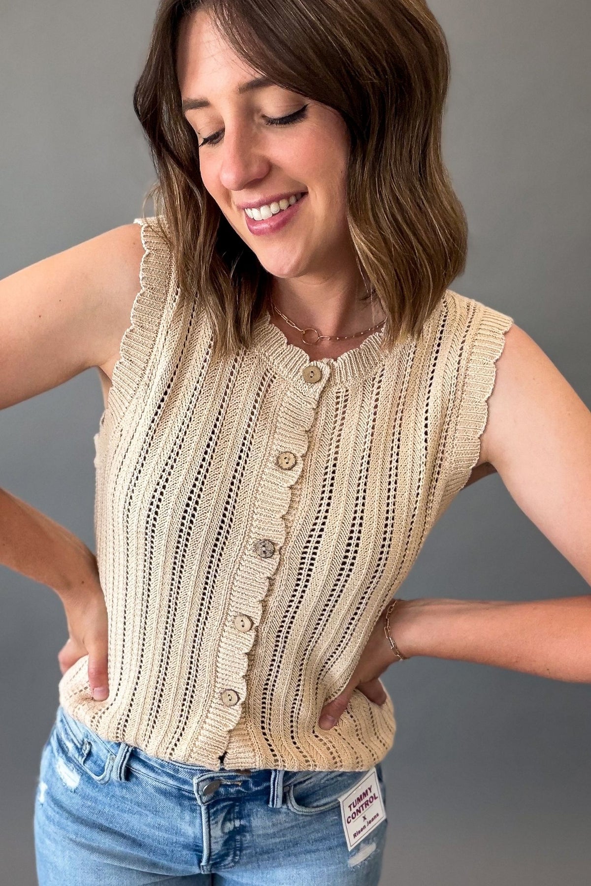 A woman wearing a sleeveless natural knit button-front top with vertical textured knit and scalloped edges, paired with blue jeans, against a plain gray background.