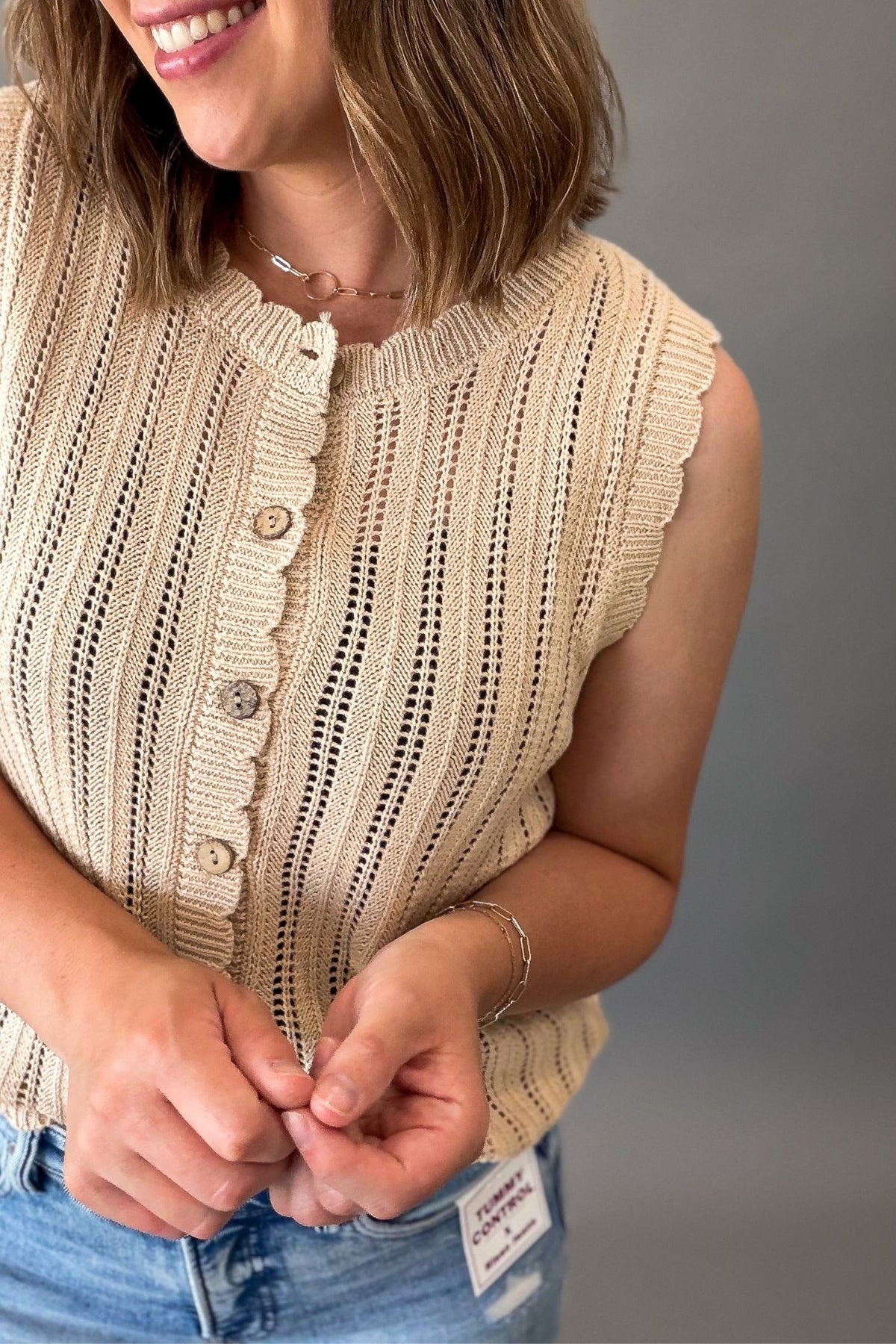 A woman wearing a sleeveless natural knit top with scalloped edges and front buttons, paired with blue jeans, shown against a plain gray background.