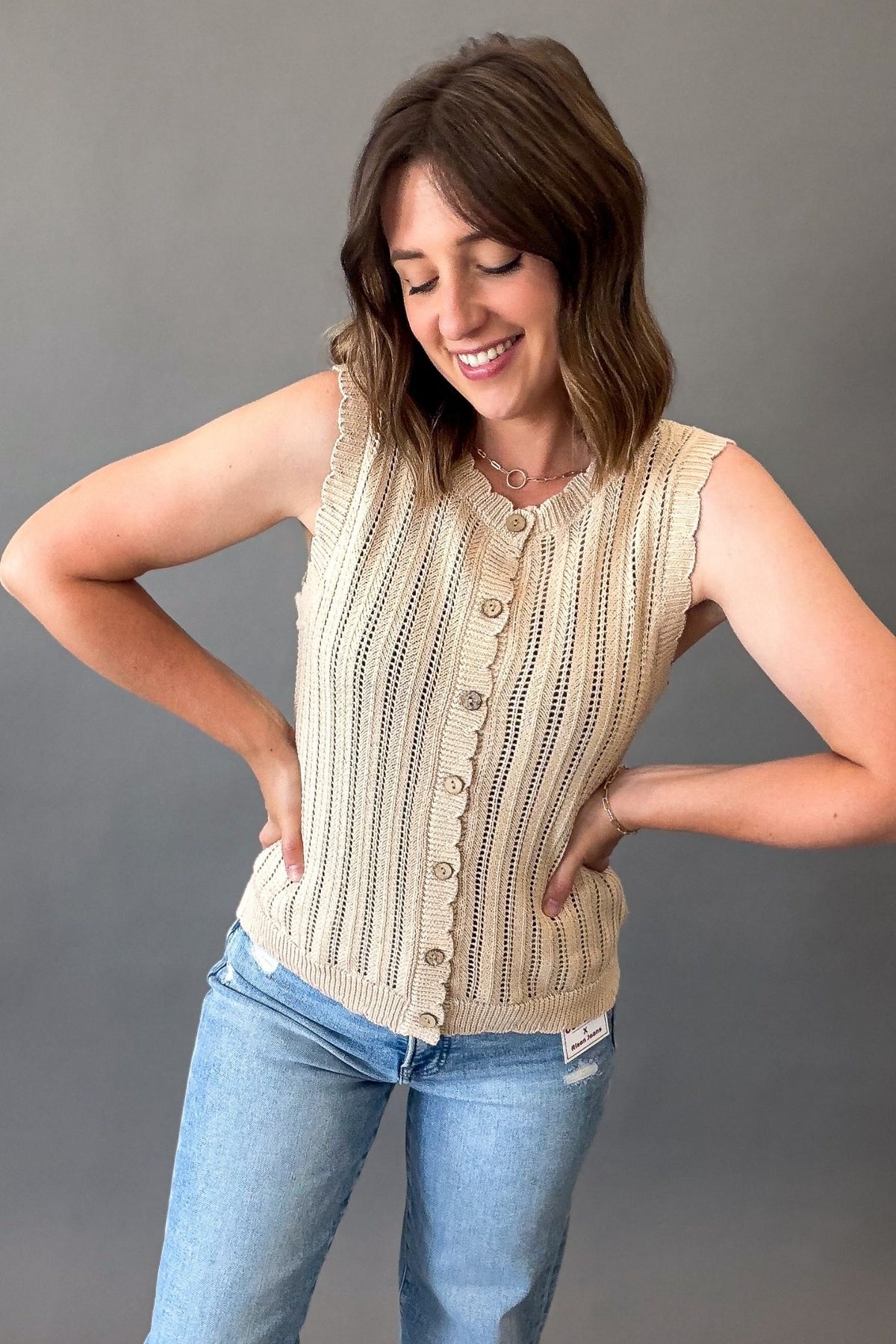 woman wearing a crochet scalloped button down tank in natural with blue jeans fully buttoned on a grey background
