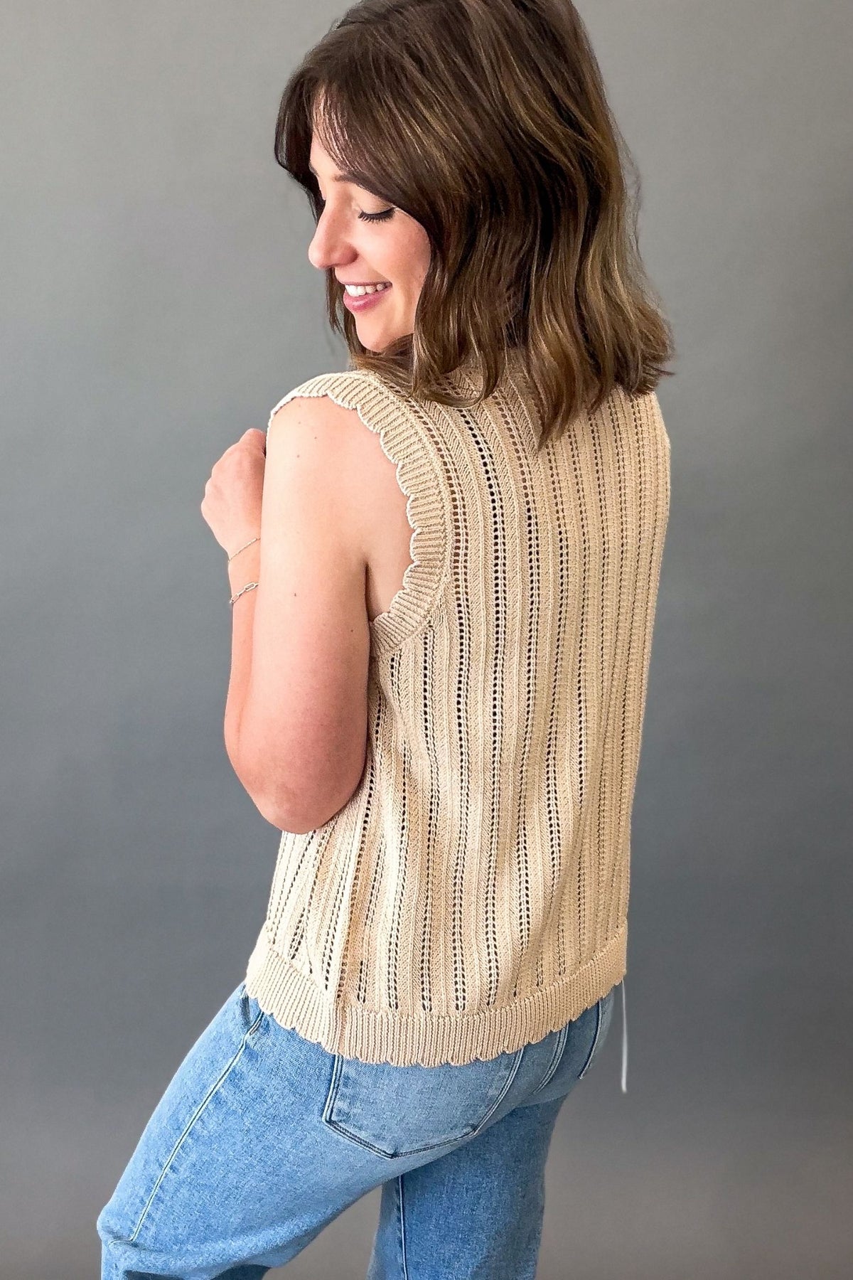A woman wearing a sleeveless beige knit top with vertical openwork detailing and scalloped edges, paired with blue jeans, photographed against a plain gray studio background.
