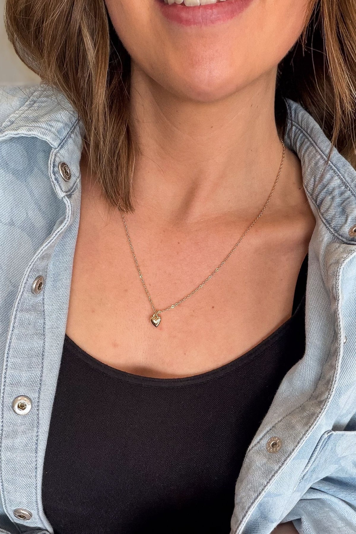 Person wearing a delicate necklace with a heart pendant, dressed in a denim jacket and black top.