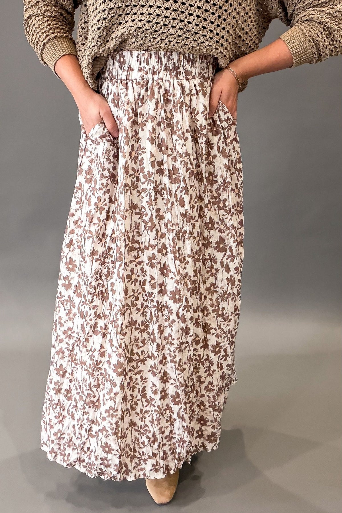 A person wearing a long pleated maxi skirt with a brown and cream floral pattern, paired with a chunky knit sweater and tan boots, shown against a neutral gray studio background.
