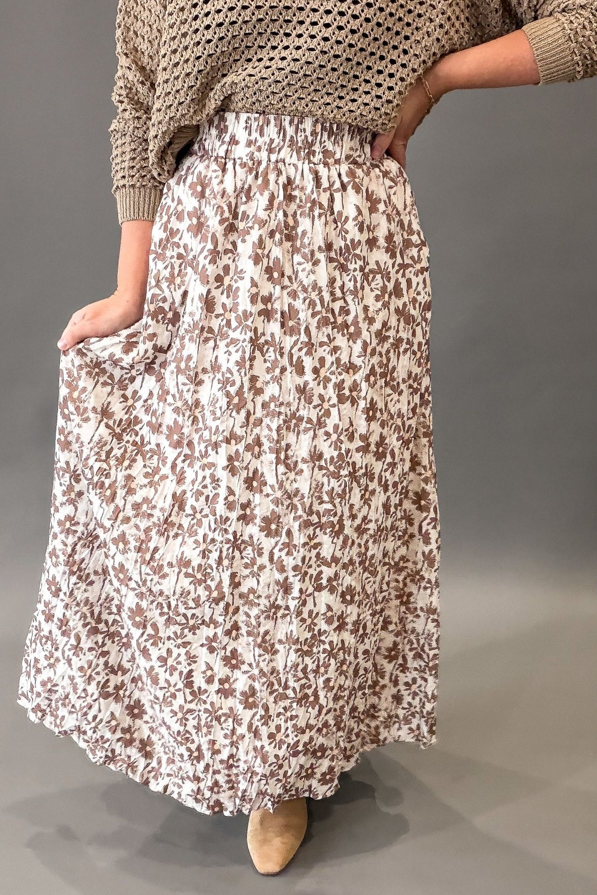 A woman wearing a long pleated maxi skirt with a brown and beige floral pattern, paired with a chunky knit beige sweater and tan boots, shown against a gray studio background.