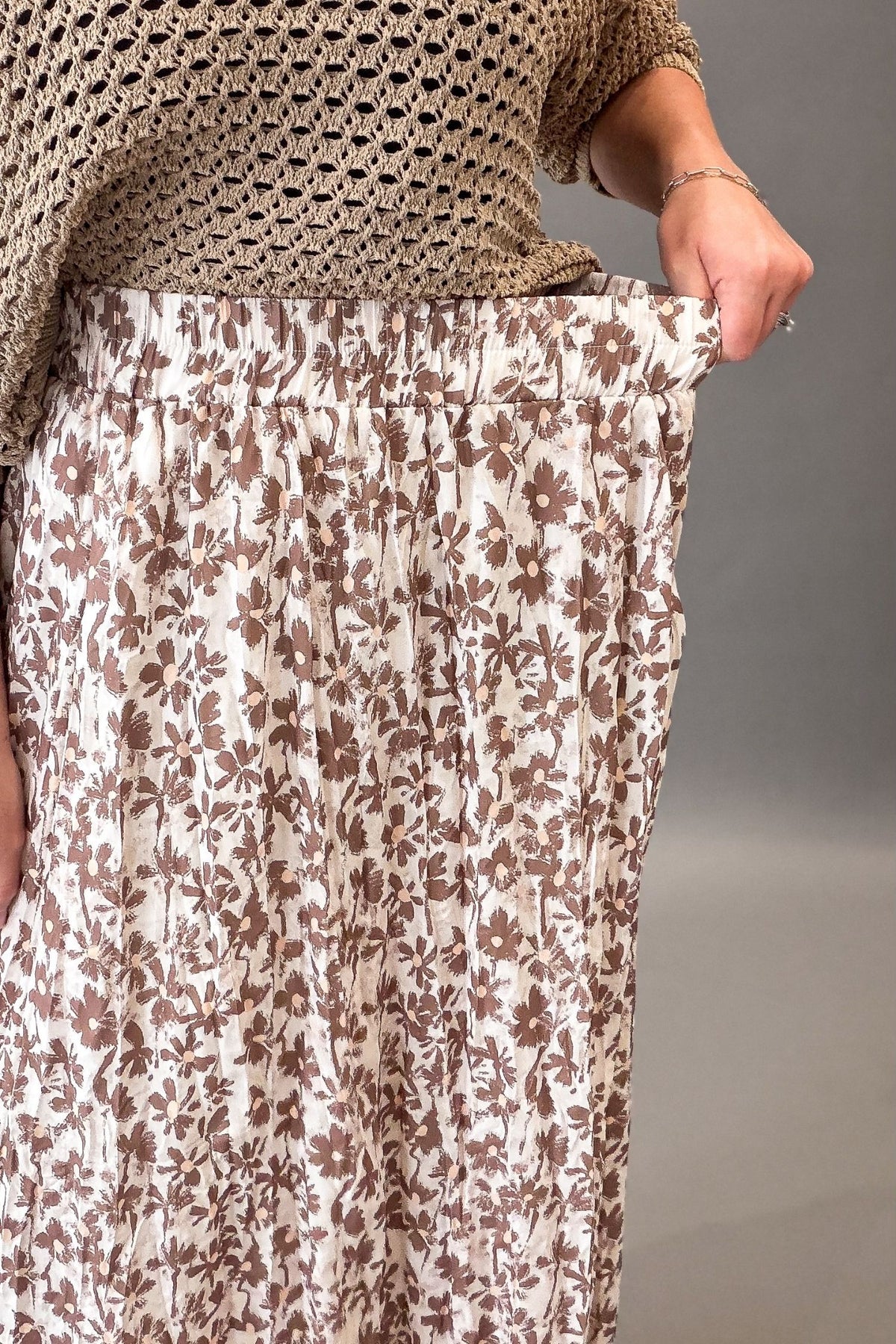 A close-up of a person wearing a cream-colored pleated skirt with a brown floral pattern, paired with a beige crochet top, against a neutral gray background.