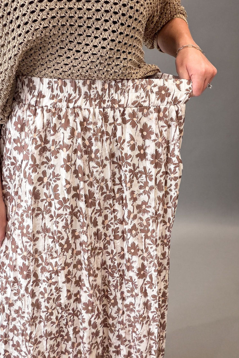 A close-up of a person wearing a cream-colored pleated skirt with a brown floral pattern, paired with a beige crochet top, against a neutral gray background.